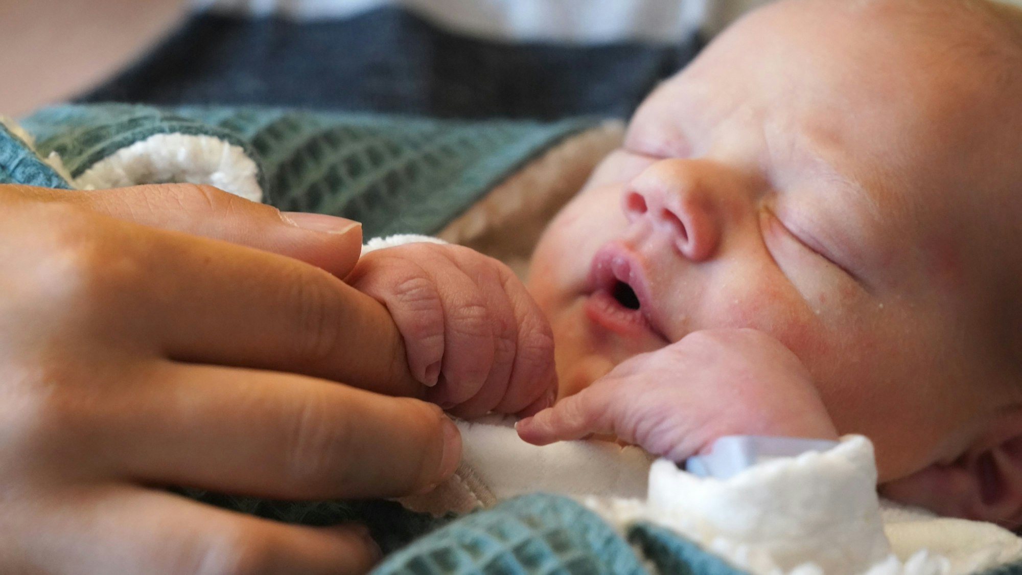 Ein Neugeborenes liegt in einer Decke eingeschlagen und hält einen Finger von einer Frau.