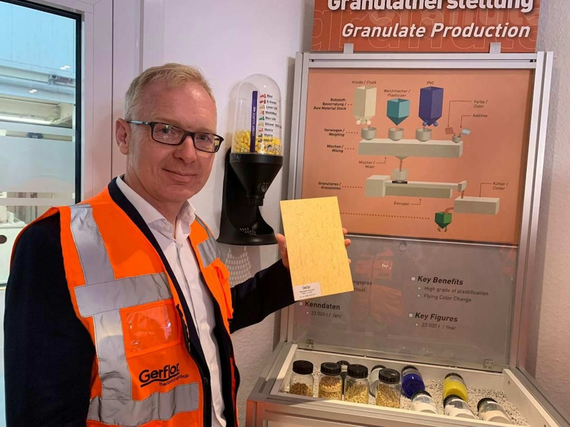 Ein Mann in einer Warnweste hält ein rechteckiges Stück PVC-Boden hoch.