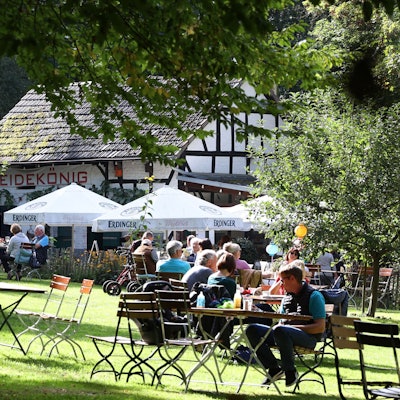 Menschen sitzen am Holztischen auf einer Wiese vor einem Fachwerkhaus