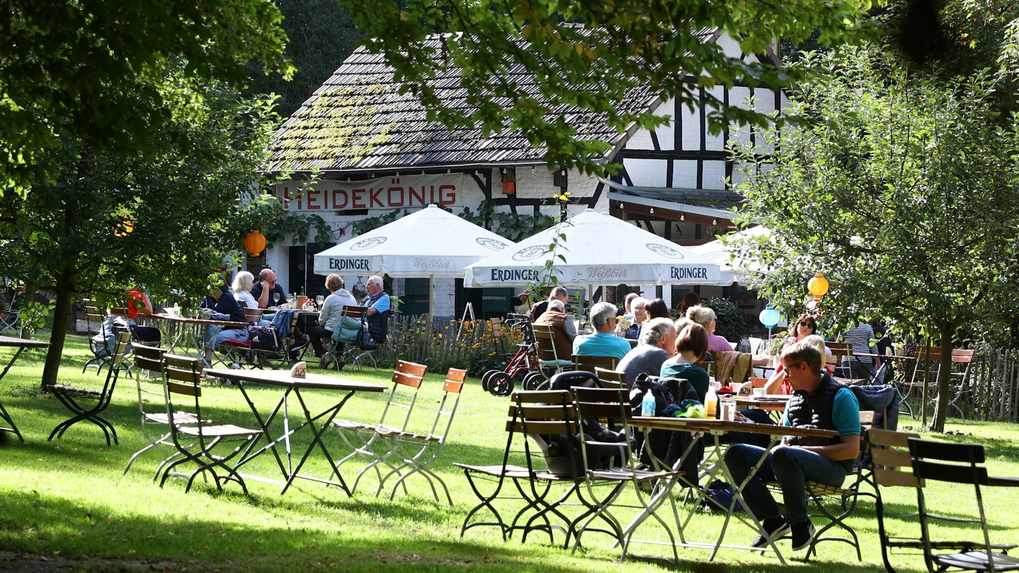 Menschen sitzen am Holztischen auf einer Wiese vor einem Fachwerkhaus