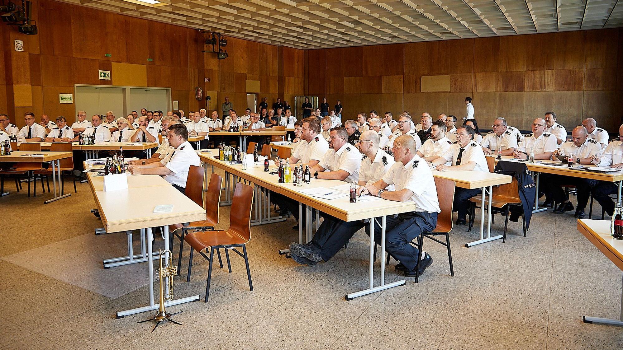 Zahlreiche Feuerwehrleute in weißen Uniformhemden sitzen in einer Halle an Tischen.