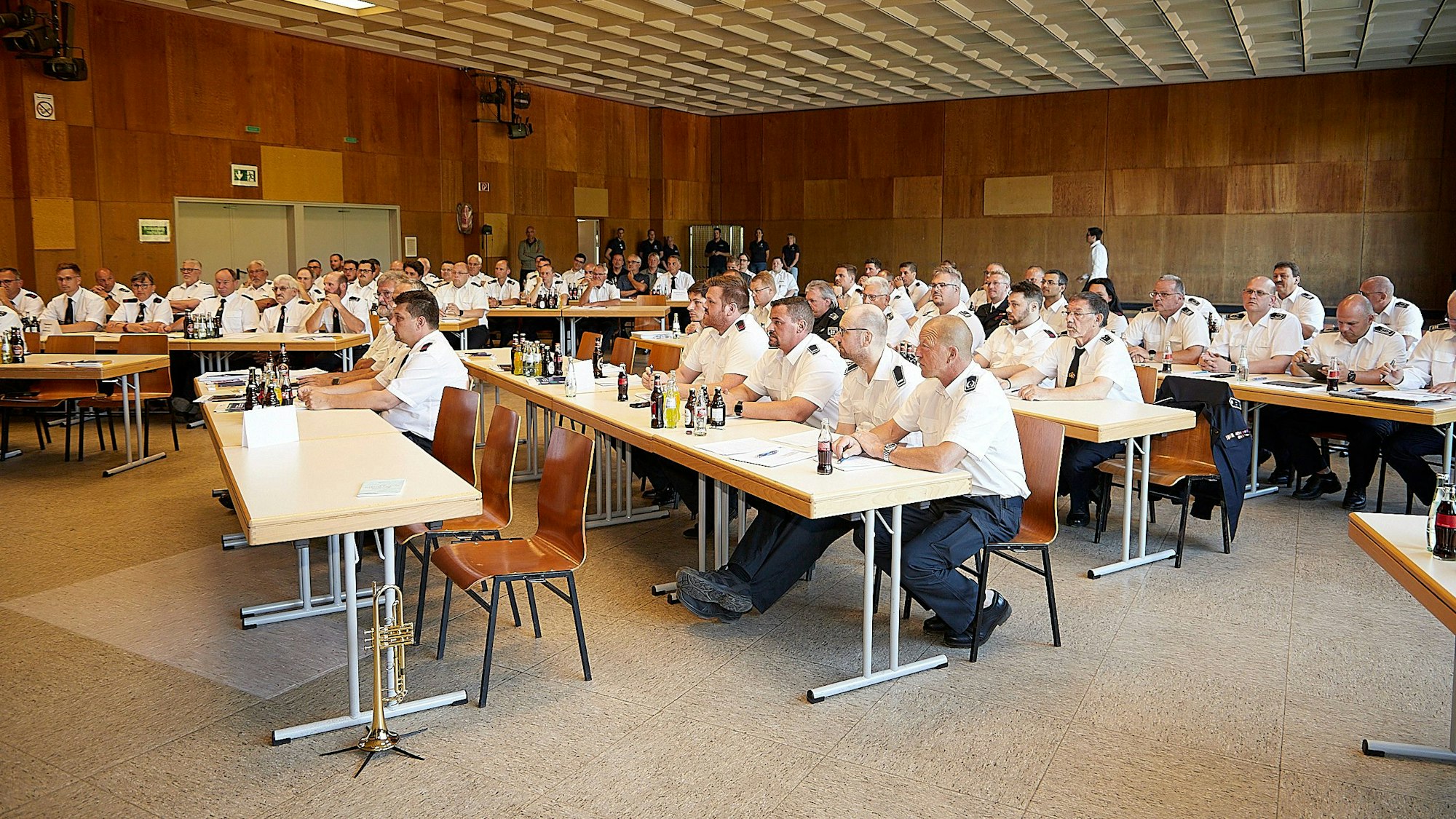 Zahlreiche Feuerwehrleute in weißen Uniformhemden sitzen in einer Halle an Tischen.