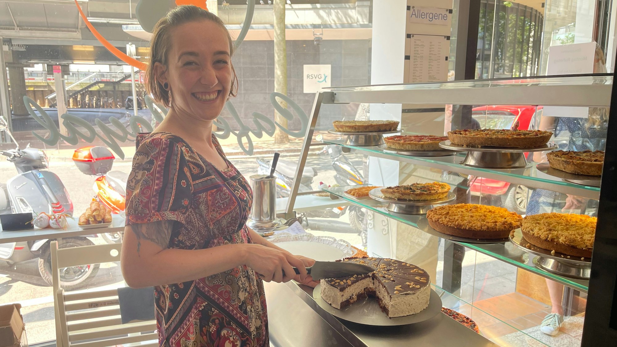 Eine Frau in einem gemusterten Kleid schneidet ein Stück Torte ab, in der Vitrine vor ihr stehen weitere Kuchen und Quiches.