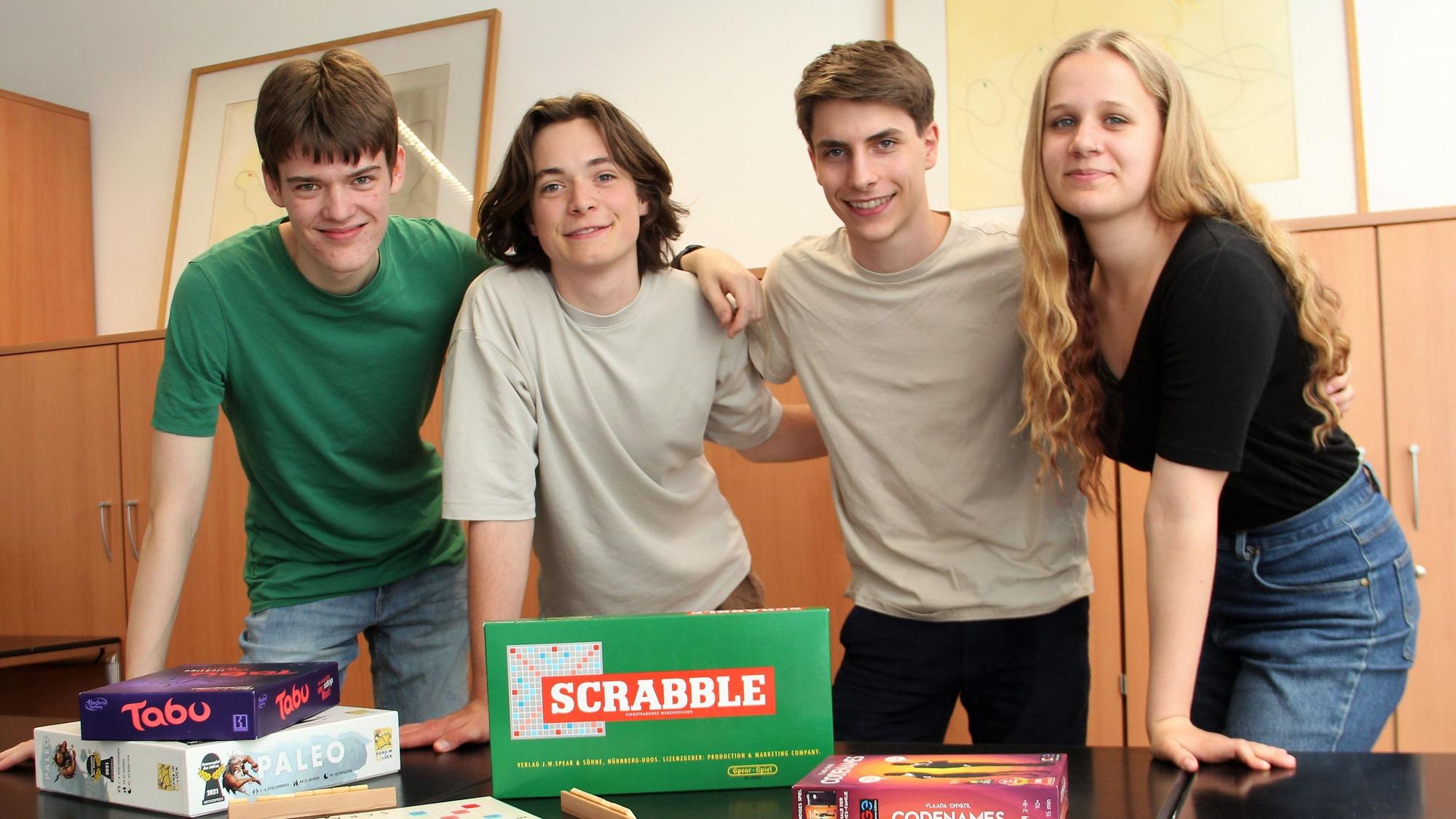 Drei junge Männer und eine junge Frau stehen an einem Tisch, auf dem Spiele, wie Tabu und Scrabble liegen.