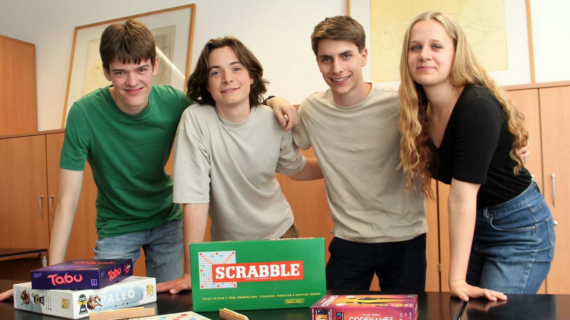 Drei junge Männer und eine junge Frau stehen an einem Tisch, auf dem Spiele, wie Tabu und Scrabble liegen.