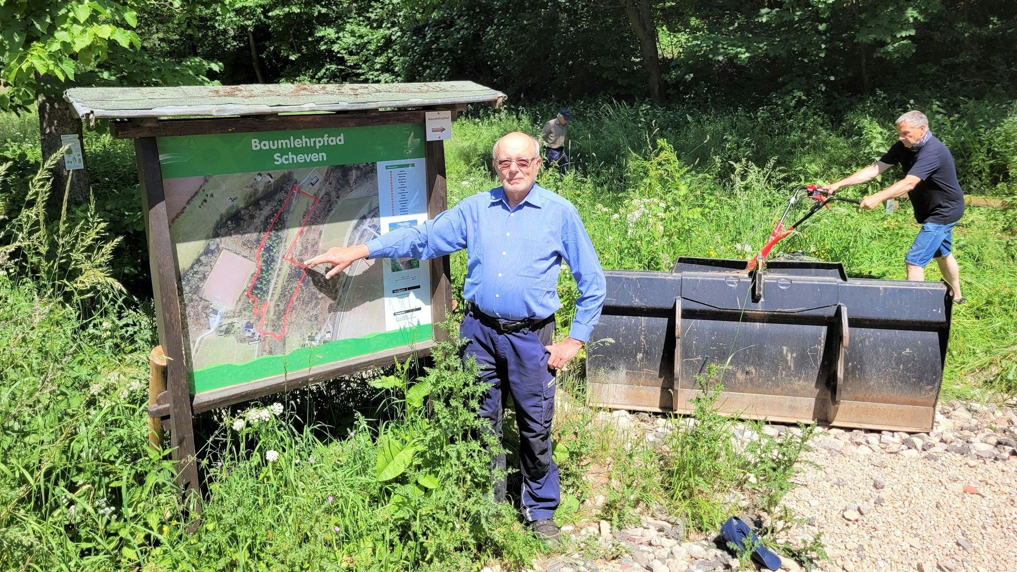 Ein Mann zeigt auf eine Informationstafel, im Hintergrund arbeitet ein weiterer Mann im Grünen.