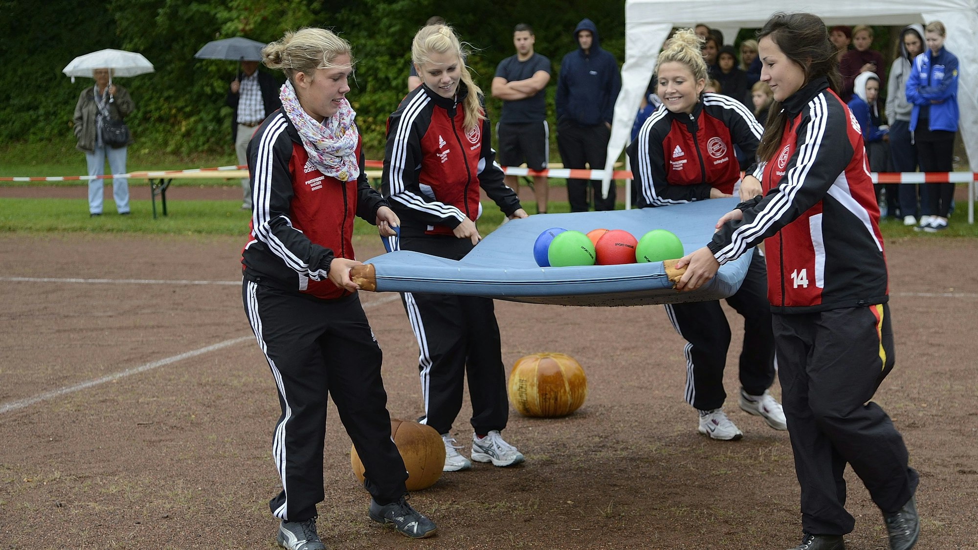 Vier Mädchen in Trainingsanzügen balancieren mehrere Bälle auf einer Turnmatte. Sie befinden sich auf einem Ascheplatz, im Hintergrund finden sich Zuschauer.