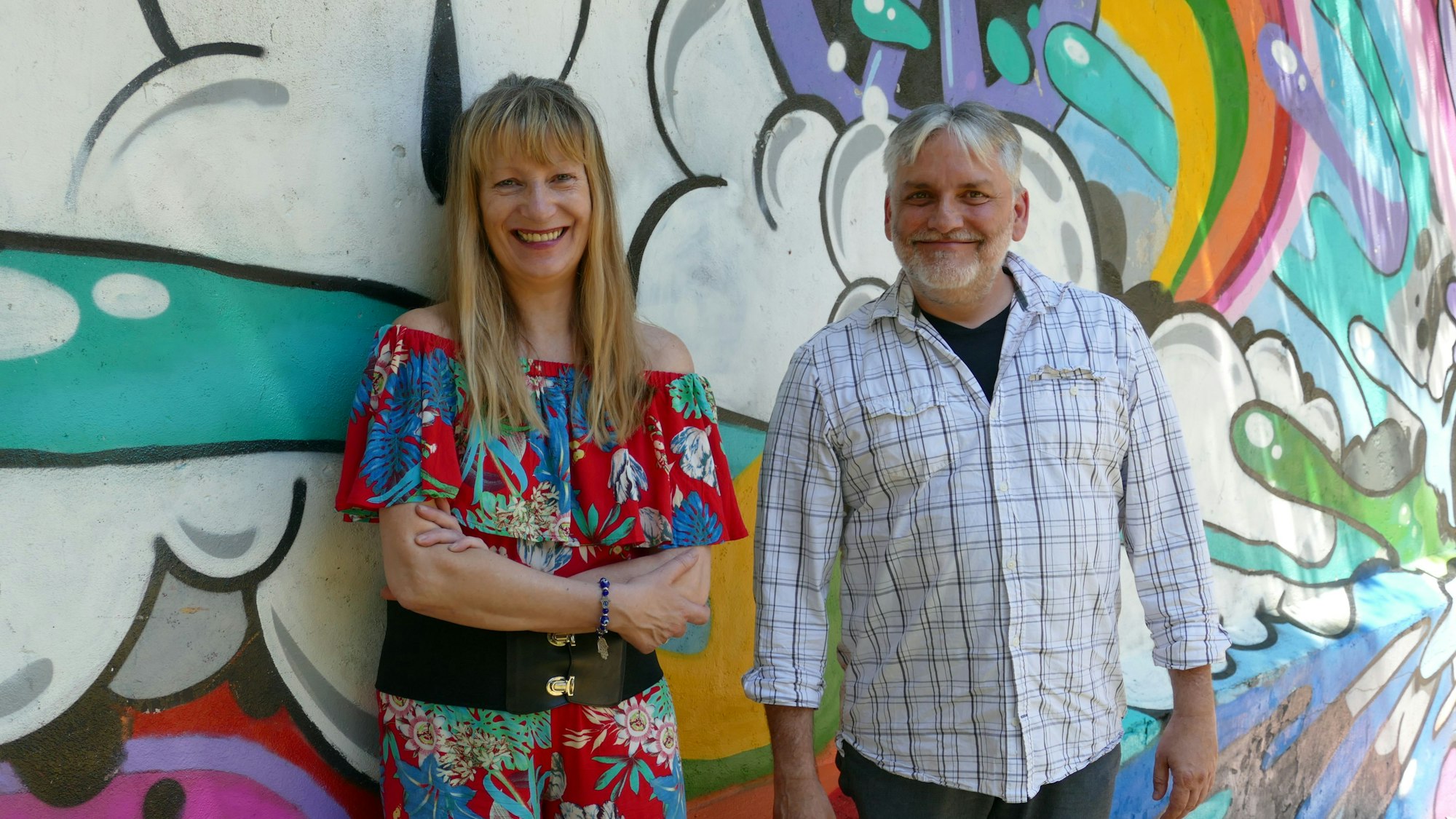 Eine Frau mit langen blonden Haaren und ein Manan stehen vor einer bunt bemalten Wand, dem Mahnmal für das Nagelbombenattentat in Mülheim.