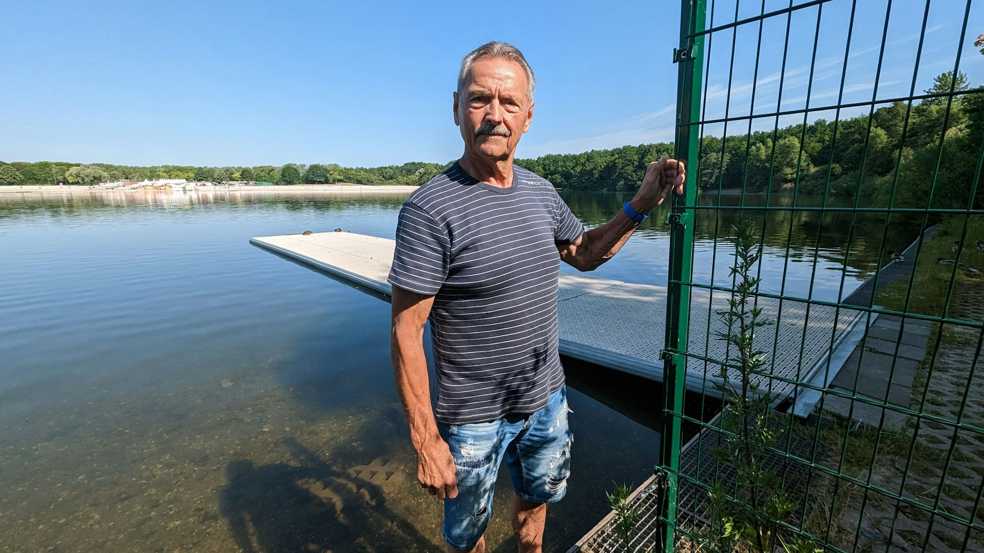 Kunze steht in kurzer Hose und T-Shirt neben dem Zaun. Im Hintergrund ist der Bootssteg zu sehen.
