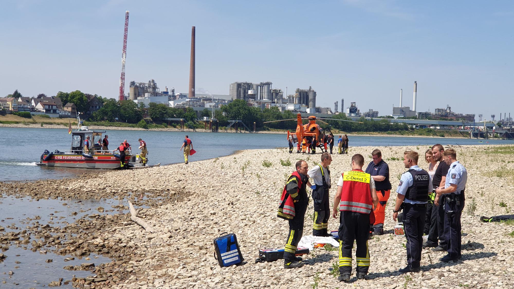 Rettungskräfte stehen am Strand des Rheins in Niederkassel. Vor dem Ufer liegt ein Boot, ein Rettungshubschrauber steht weiter hinten.