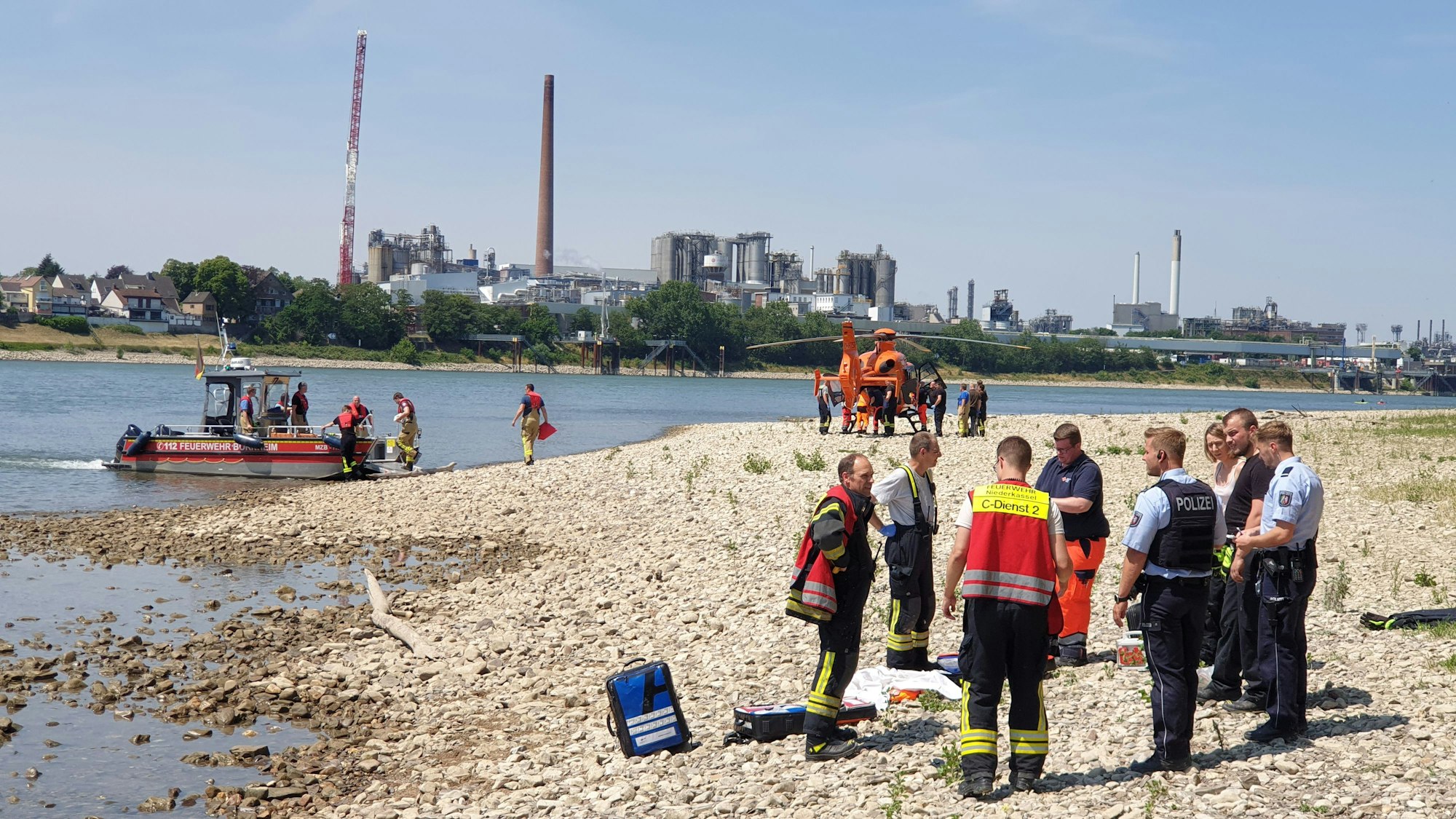Rettungskräfte stehen am Strand des Rheins in Niederkassel. Vor dem Ufer liegt ein Boot, ein Rettungshubschrauber steht weiter hinten.