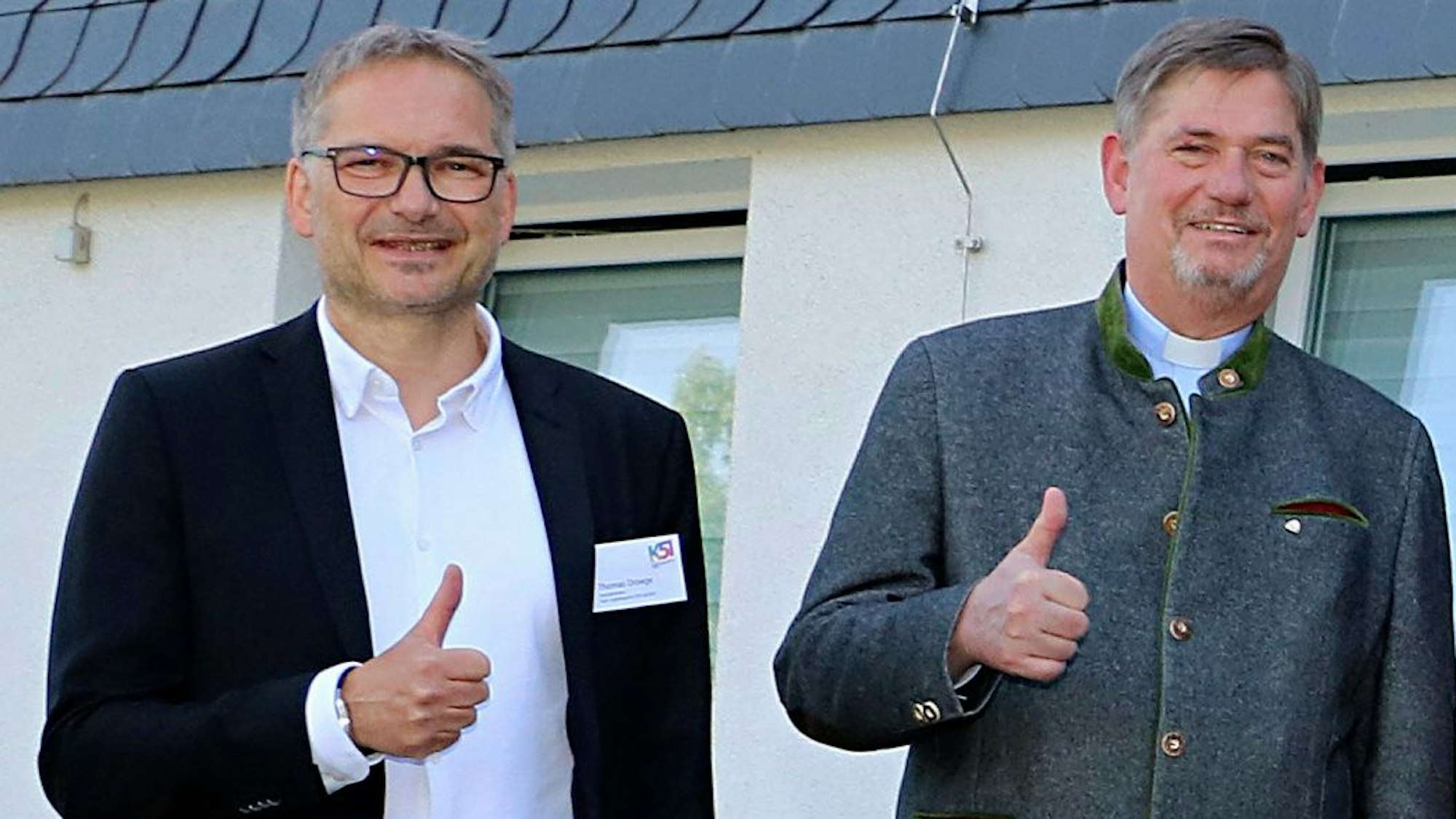 Zwei Männer stehen mit nach oben gestrecktem Daumen vor einem Gebäude.