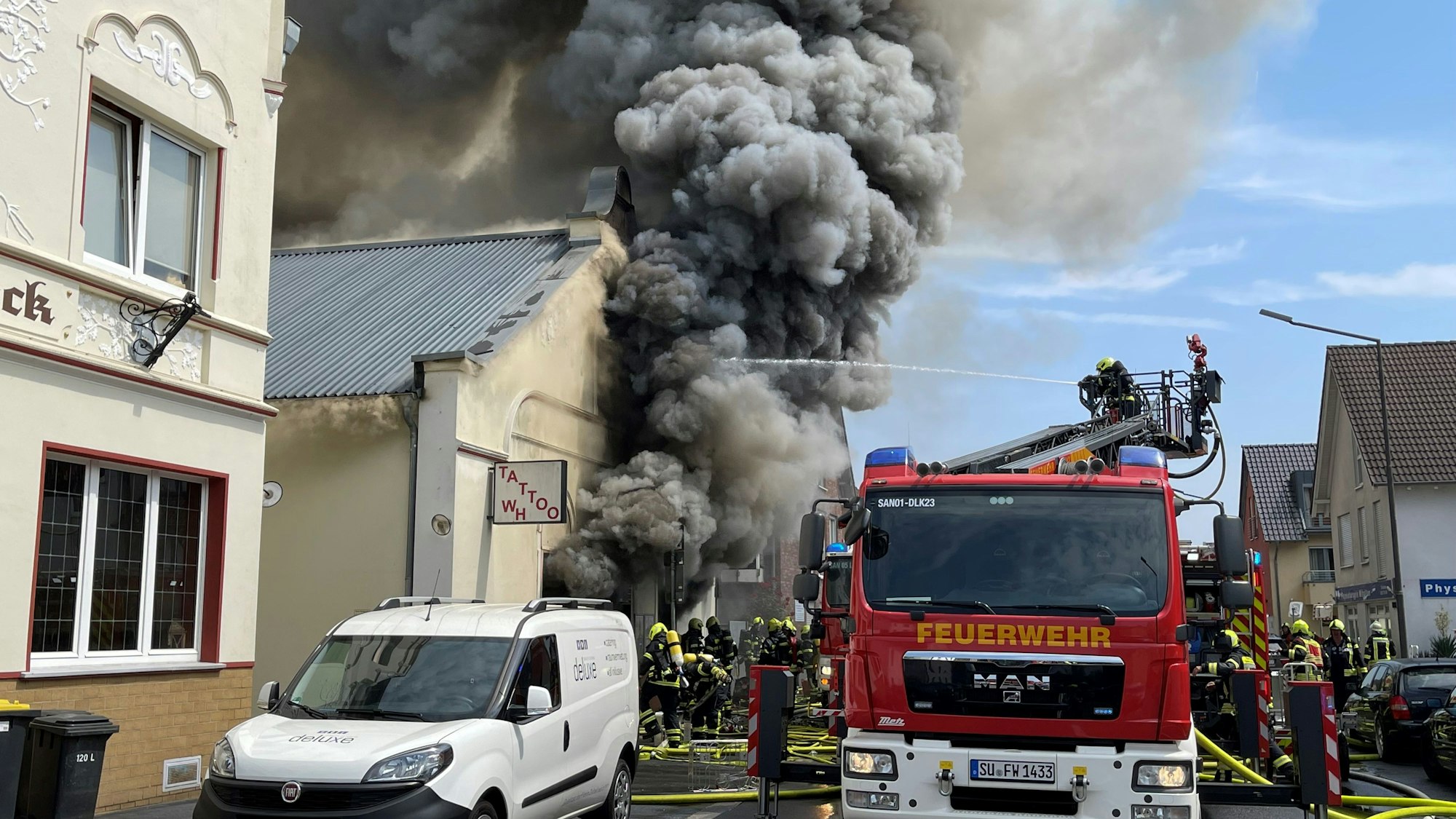 Aus einem weißen Giebelgebäude dringt dicker, schwarzer Qualm, eine Drehleiter der Feuerwehr steht davor, viele Feuerwehrleute sind im Einsatz.