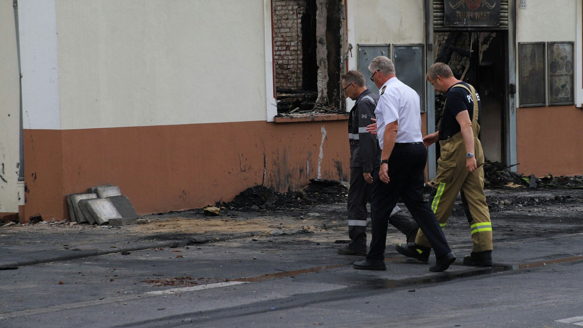 Drei Männer in Funktionskleidung gehen mit gesenkten Blicken auf das ausgebrannte Haus in Sankt Augustin zu.
