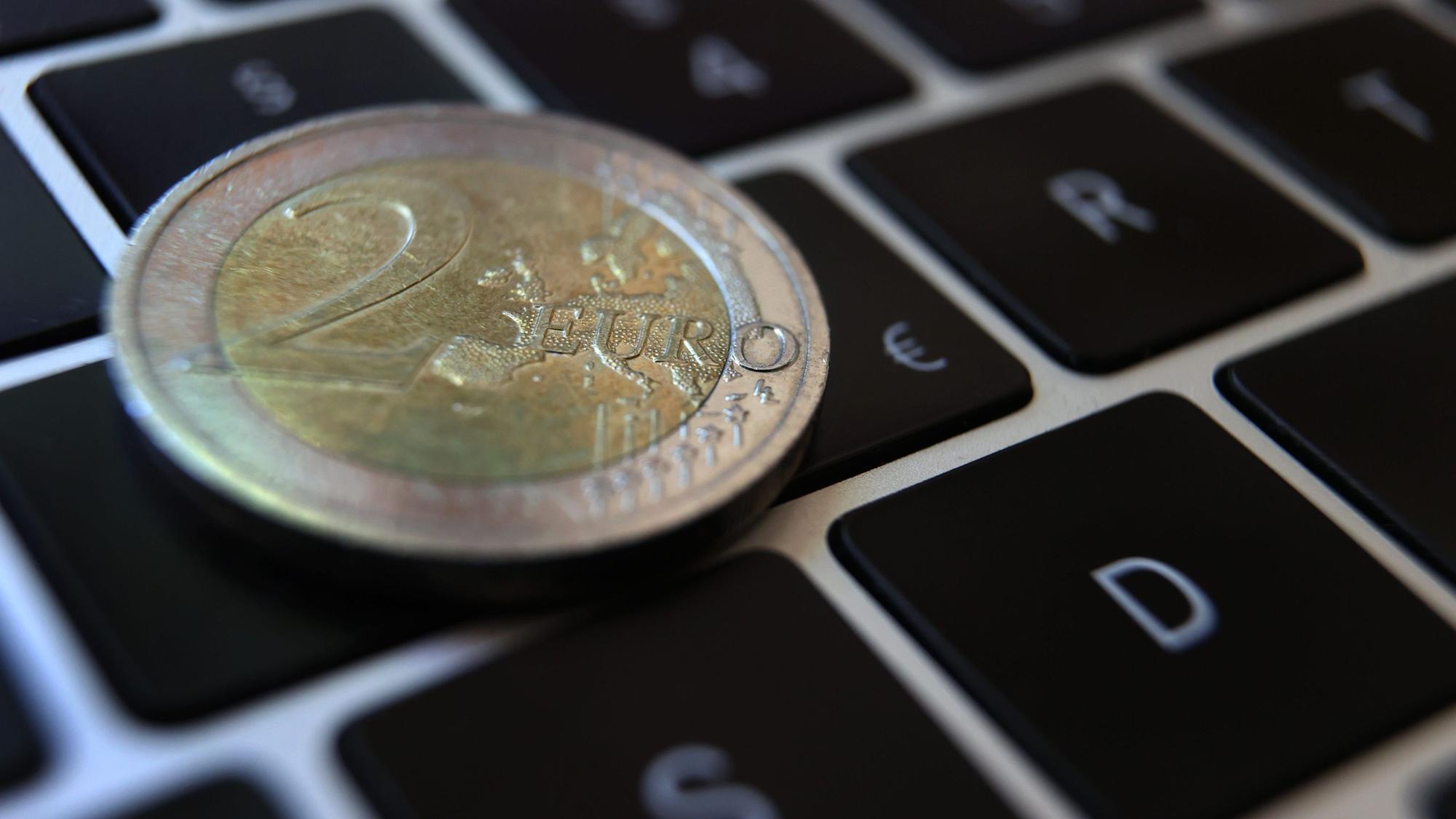 Eine Zwei -Euro-Münze liegt auf der Tastatur eines Laptops neben einem Eurozeichen.