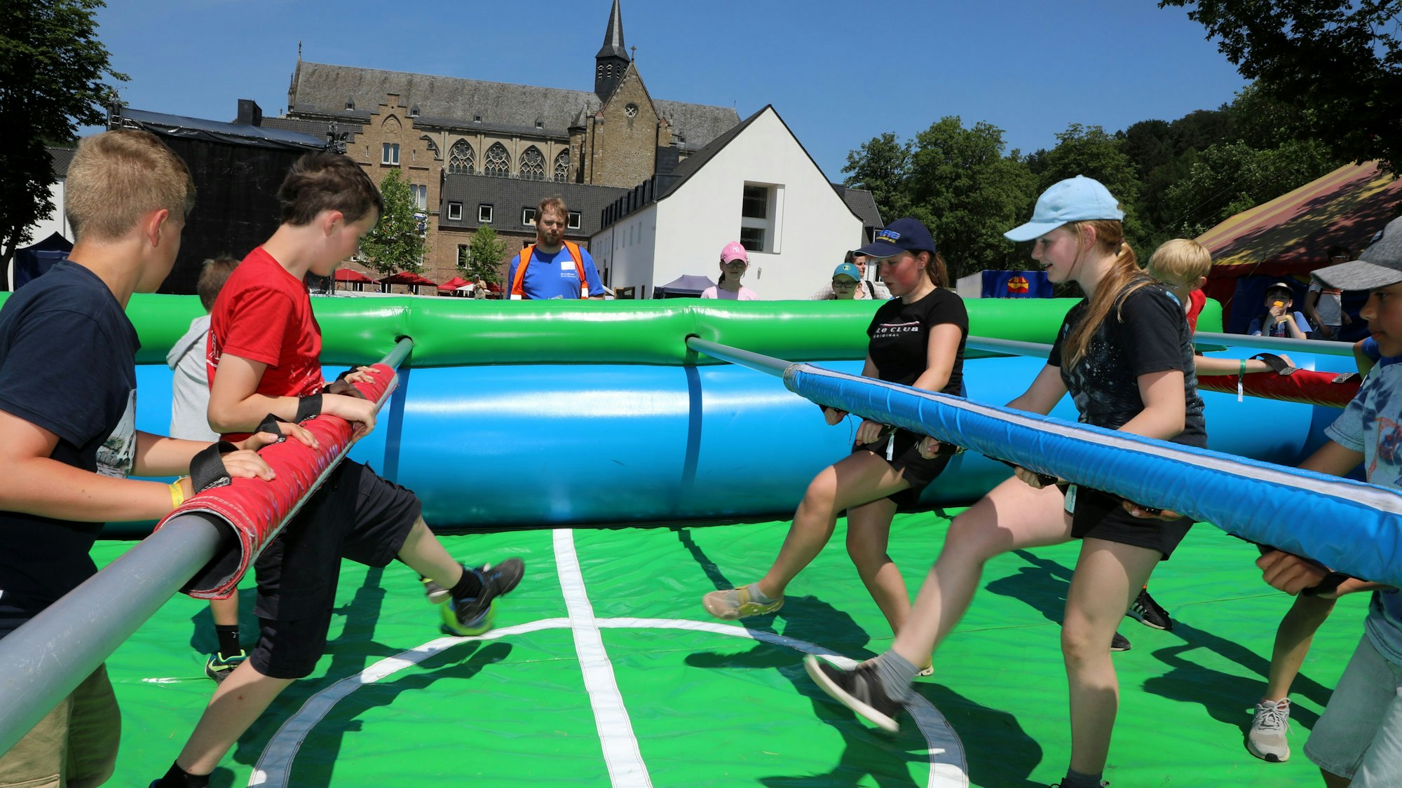 Kinder spielen an Stangen Fußball auf einem von aufblasbaren Polstern umgebenen Kleinfußballfeld. Im Hintergrund: Der Altenberger Dom.