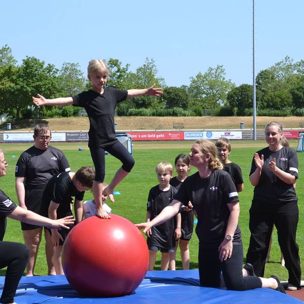 Eine junge Teilnehmerin balanciert auf einem Gymnastikball, mehrere Akteure unterstützen sie dabei.