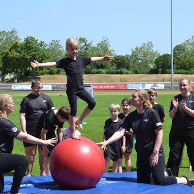 Eine junge Teilnehmerin balanciert auf einem Gymnastikball, mehrere Akteure unterstützen sie dabei.