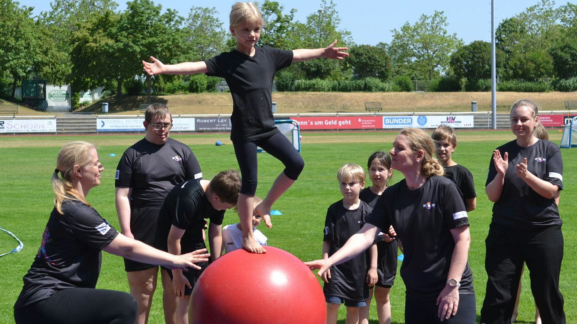 Eine junge Teilnehmerin balanciert auf einem Gymnastikball, mehrere Akteure unterstützen sie dabei.