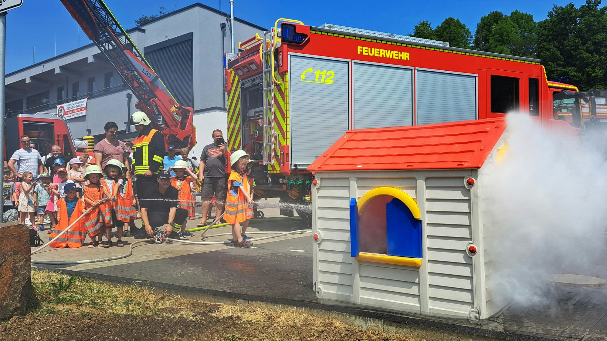 Rauch kommt aus einem Kinderspielhaus, Kinder löschen mit großen Helmen, Warnwesten und kleinen Feuerwehrschläuchen.