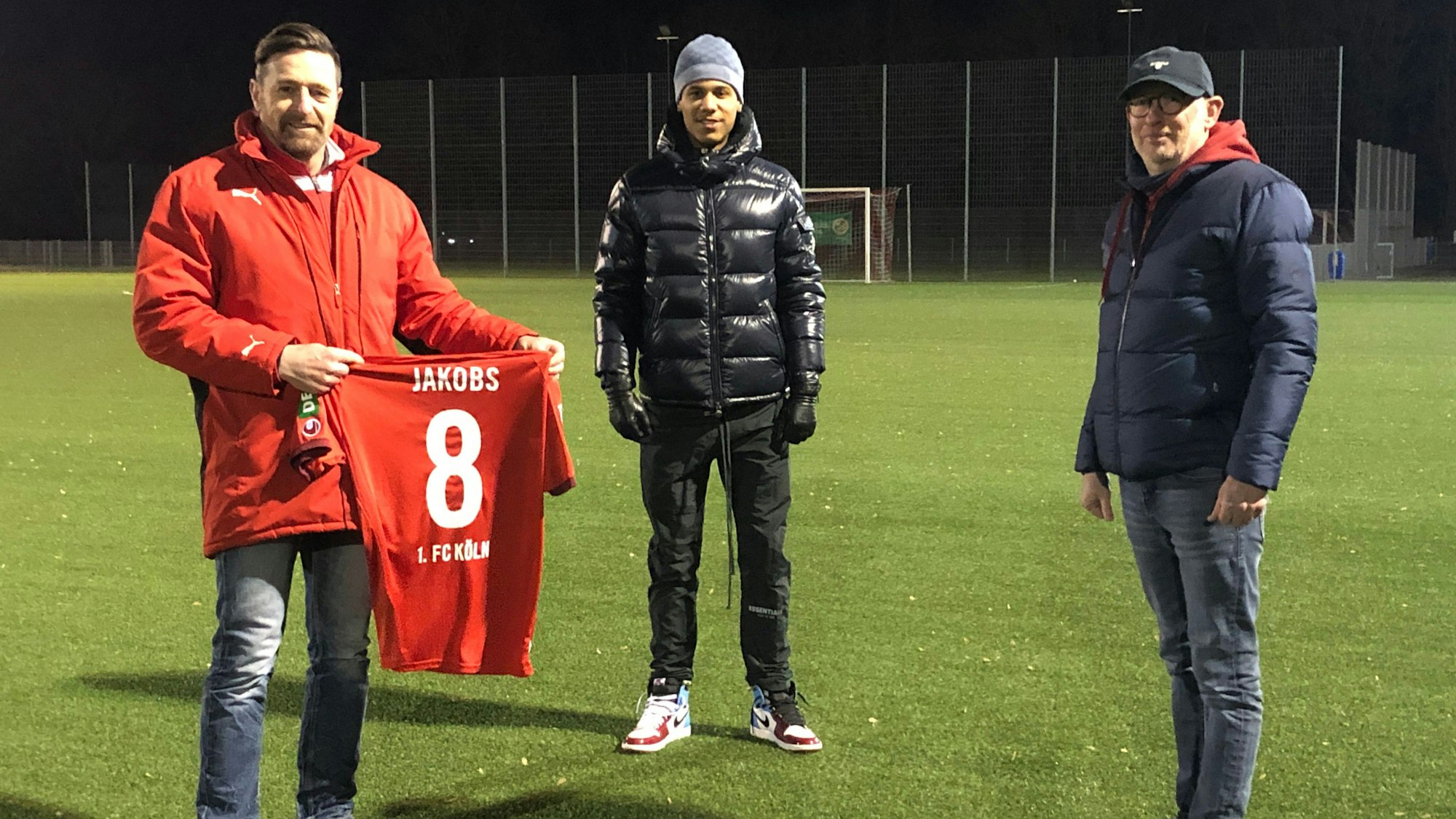 Ismail Jakobs (M.), Jugendleiter Andreas Haepp (l.) und Bliesheims Vorsitzender Alfred Schwanke schauen in die Kamera und stehen auf einem Rasenplatz. Andreas Haepp hält ein Trikot mit der Aufschrift „Jakobs“ ins Bild.
