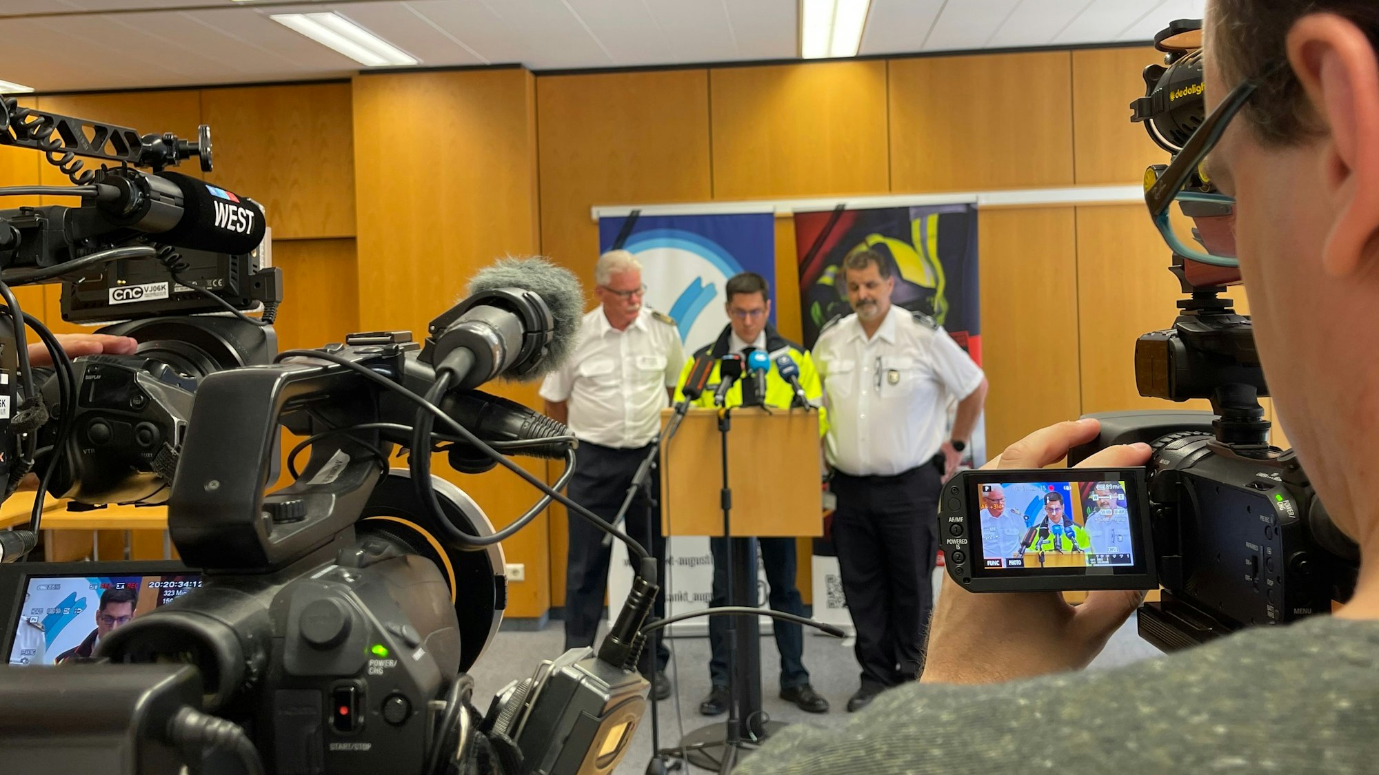 Drei Männer stehen an einem Rednerpult, Kameras und Mikrofone sind auf sie gerichtet