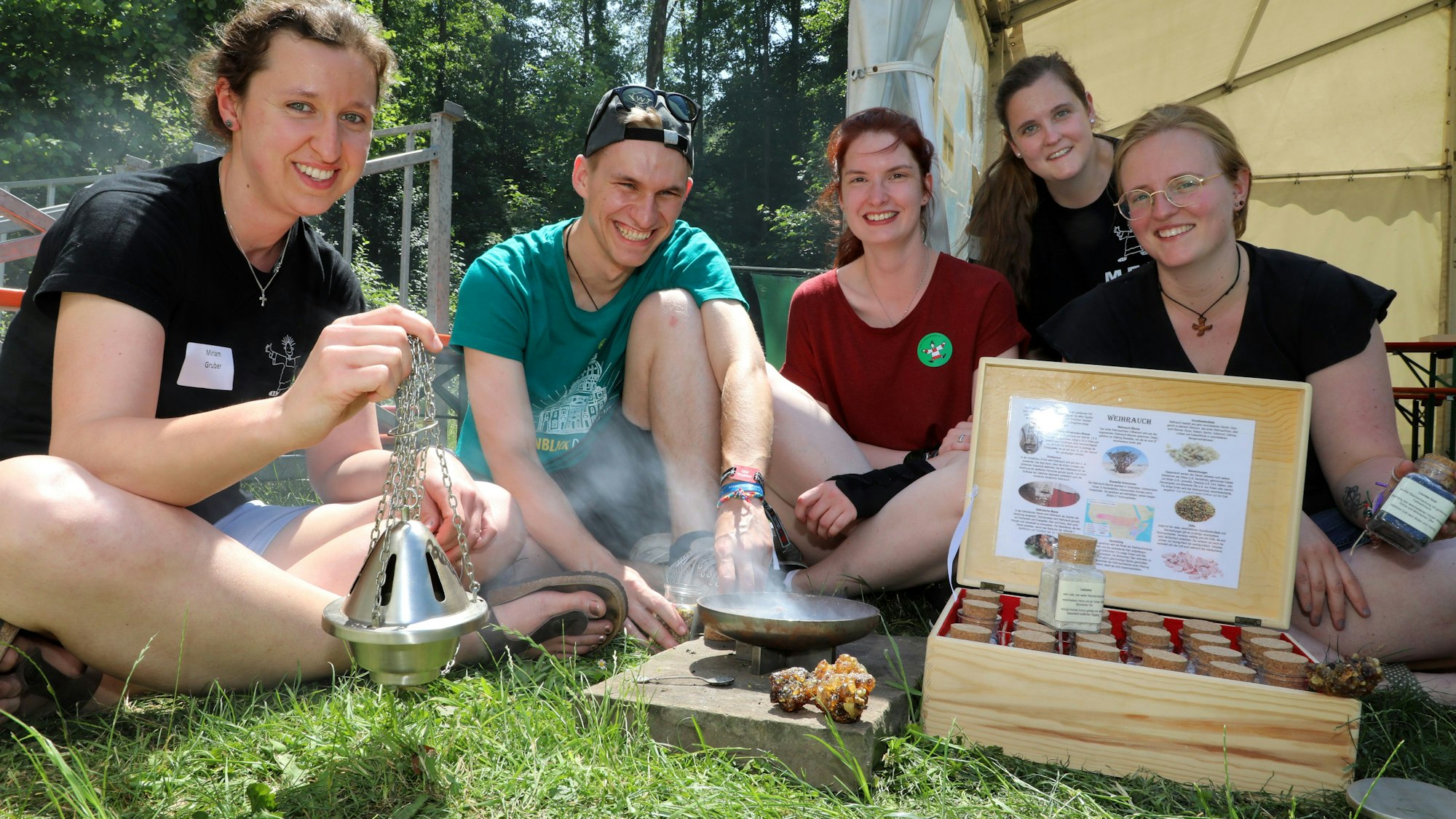 Junge Erwachsene sitzen mit Weihrauchfass, einer Weihrauchauswahl und einem Kohlefeuerchen auf einer Wiese vor einem Zelt.