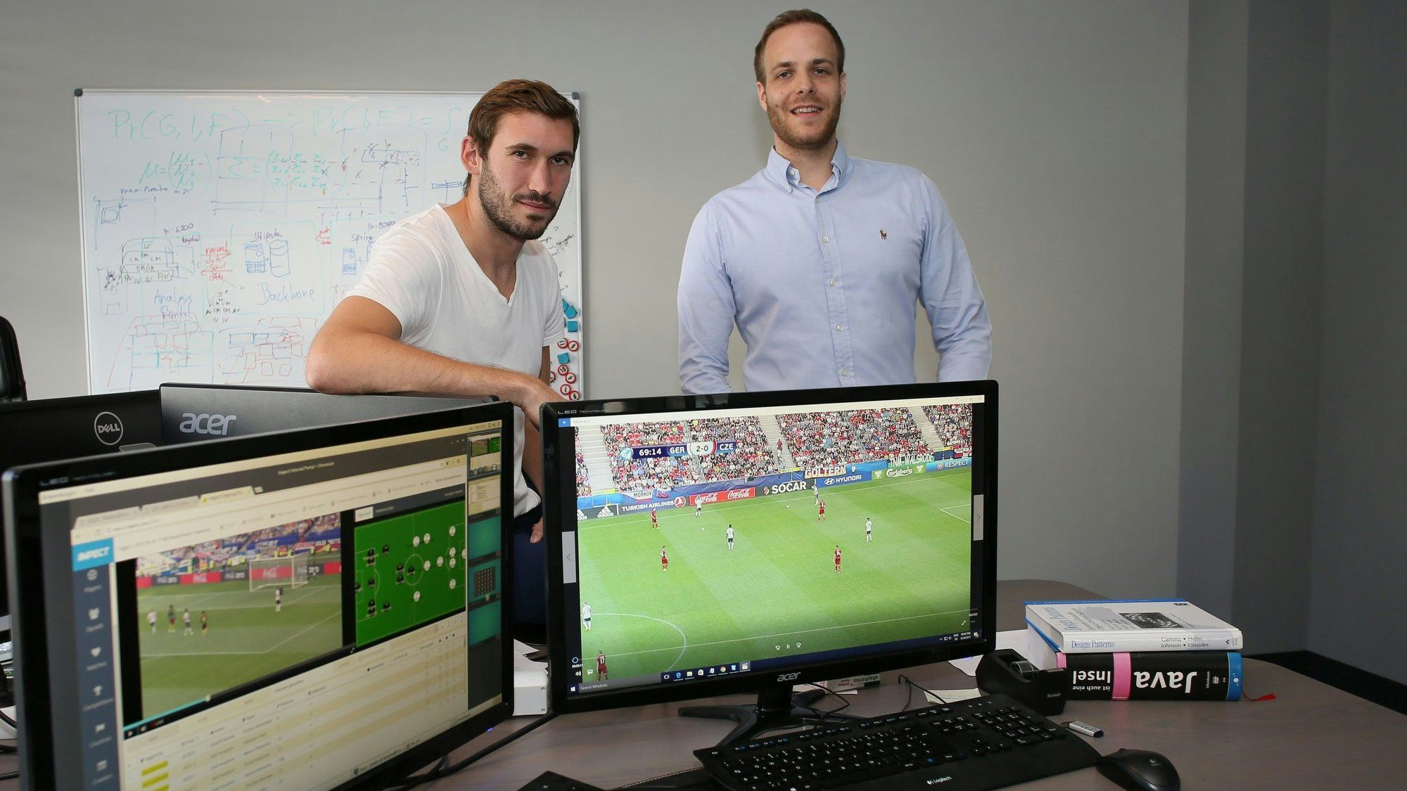 Der Ex-Nationalspieler Stefan Reinartz (li.) und Matthias Sienz (r.) schauen in die Kamera. Vor ihnen sind zwei Bildschirme auf einem Schreibtisch samt Tastaturen platziert, die Sequenzen aus Fußballspielen zeigen.