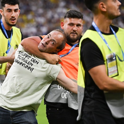 Eine Person, die auf das Spielfeld rannte und ein T-Shirt mit der Aufschrift „Stoppt den fossilen Wahnsinn!“ trägt, wird von Ordnern vom Feld geführt.