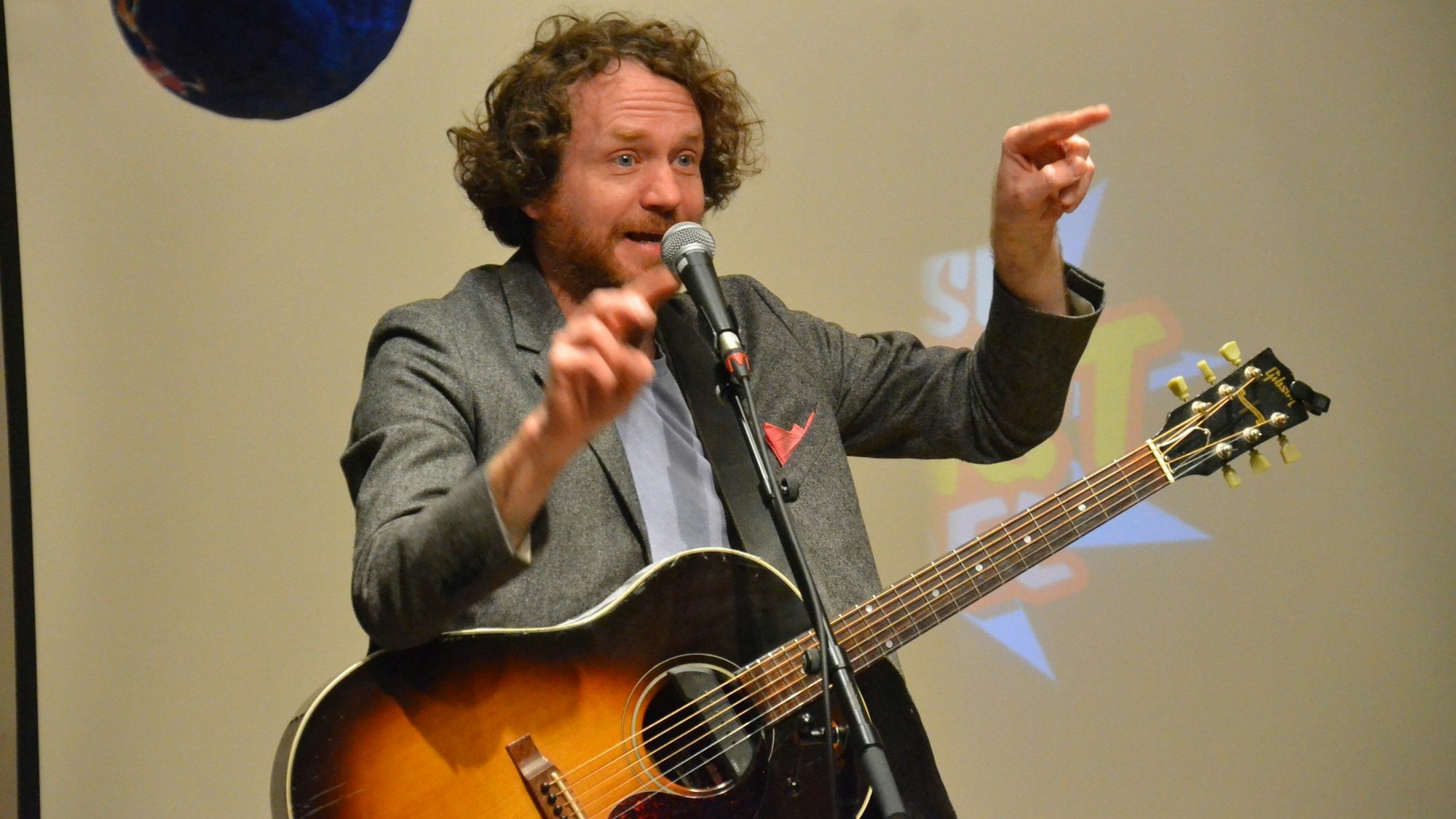 Kabarettist und Comedian Friedemann Weise steht mit einer Gitarre auf der Bühne vor einem Mikrofon und gestikuliert.