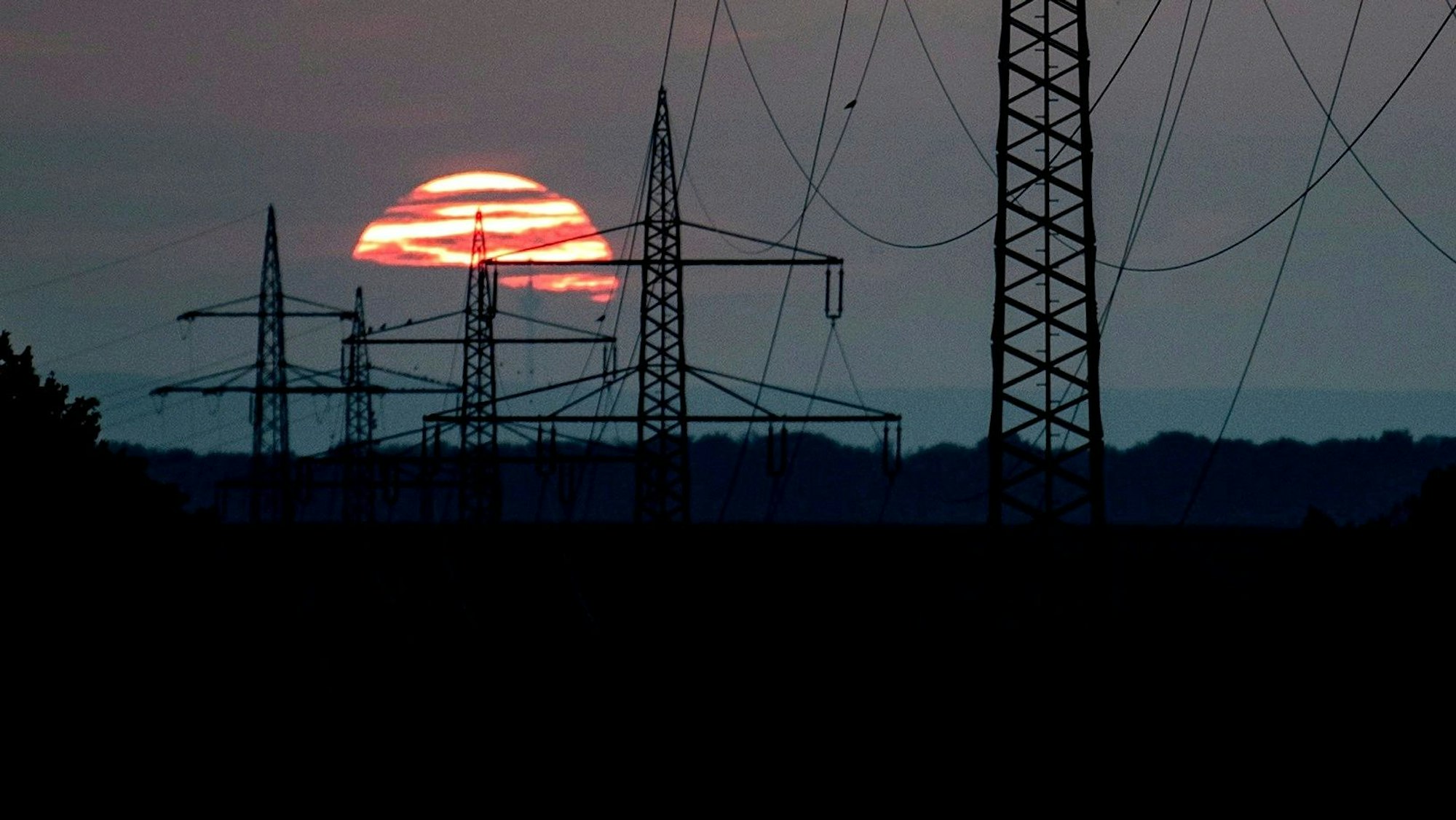 Das Bild zeigt mehrere Strommaste. Im Hintergrund geht die Sonne unter.
