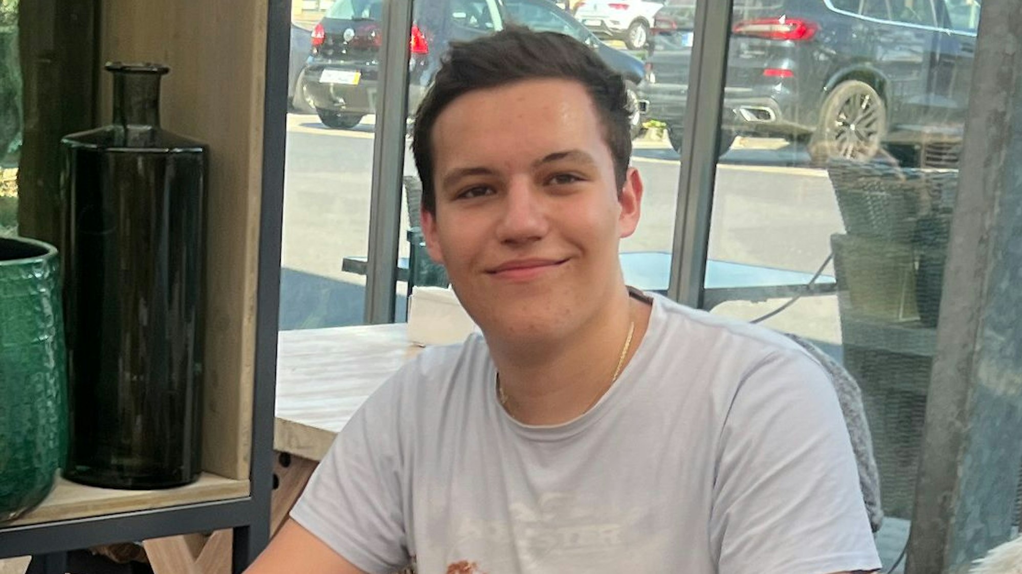 Das Porträtbild zeigt den Abiturienten Leon Tzikas. Er trägt ein weißes T-Shirt und hat kurze, braune Haare.