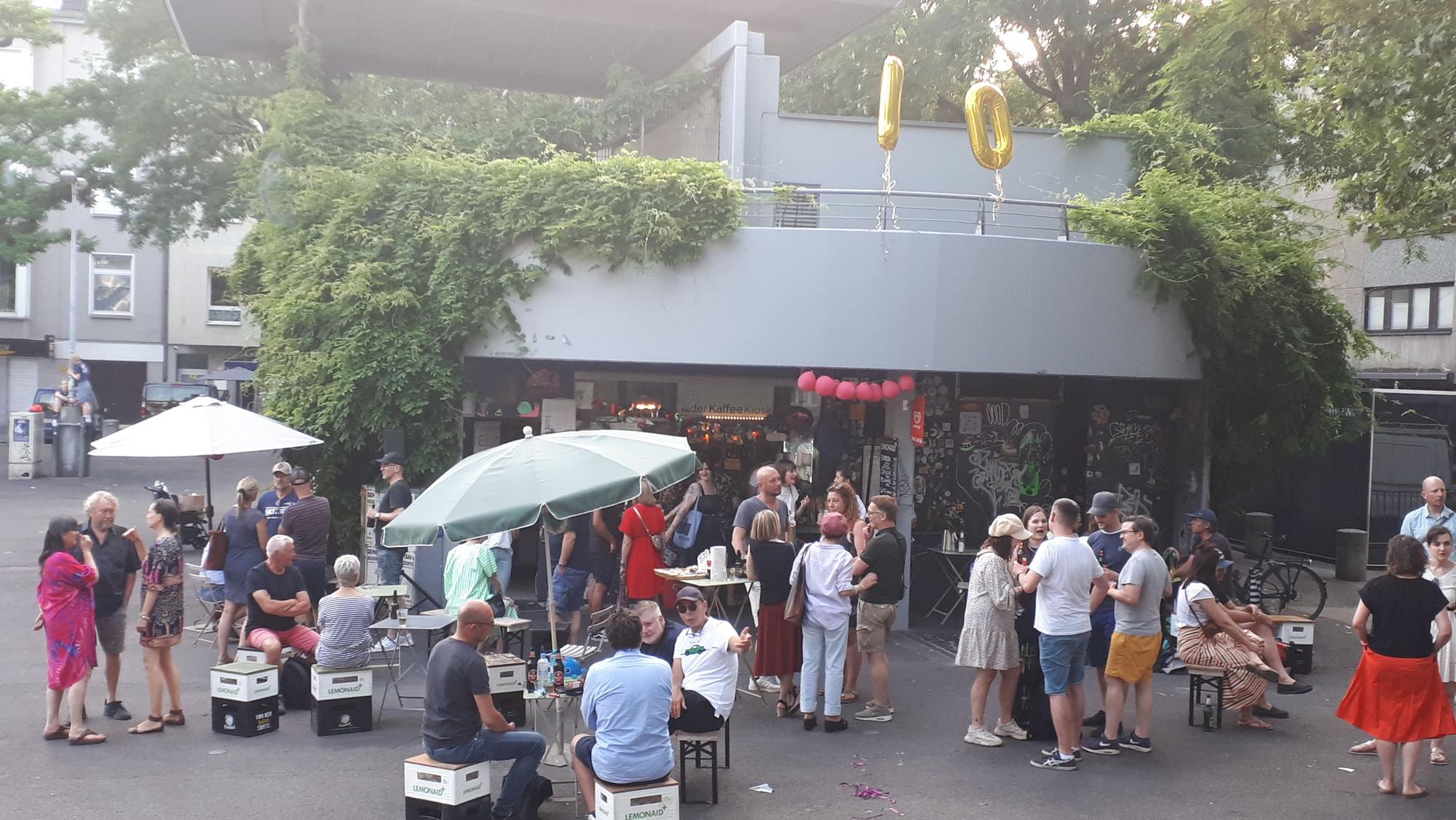 Viele Menschen stehen oder sitzen auf Hockern auf einem Platz vor einem Kiosk, der in einem Betongebäude untergebracht ist.