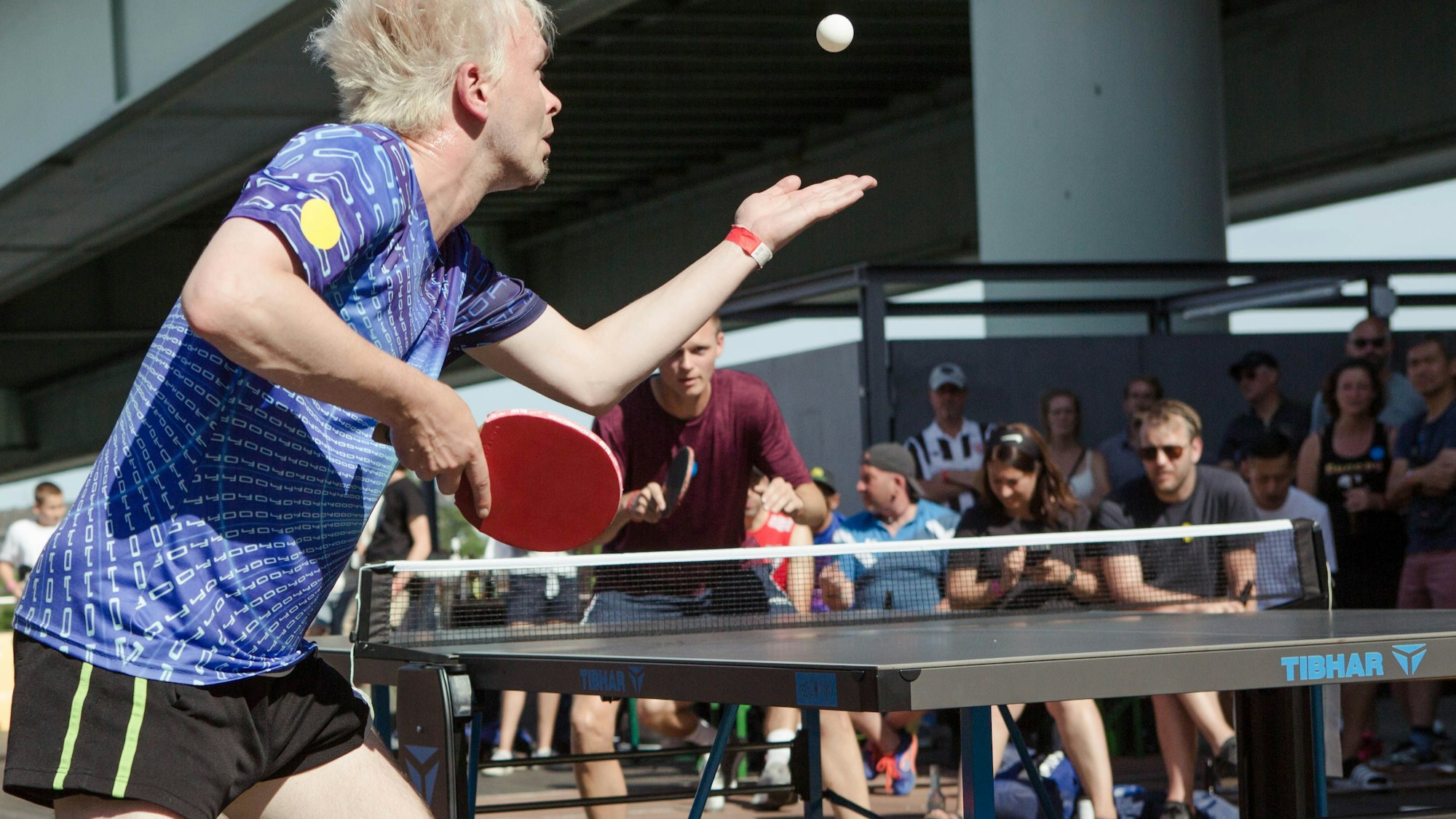 Zwei Tischtennisspieler sind mitten in einem Ballwechsel, die Platte steht draußen auf Holzboden, im Hintergrund sind Zuschauer zu sehen.