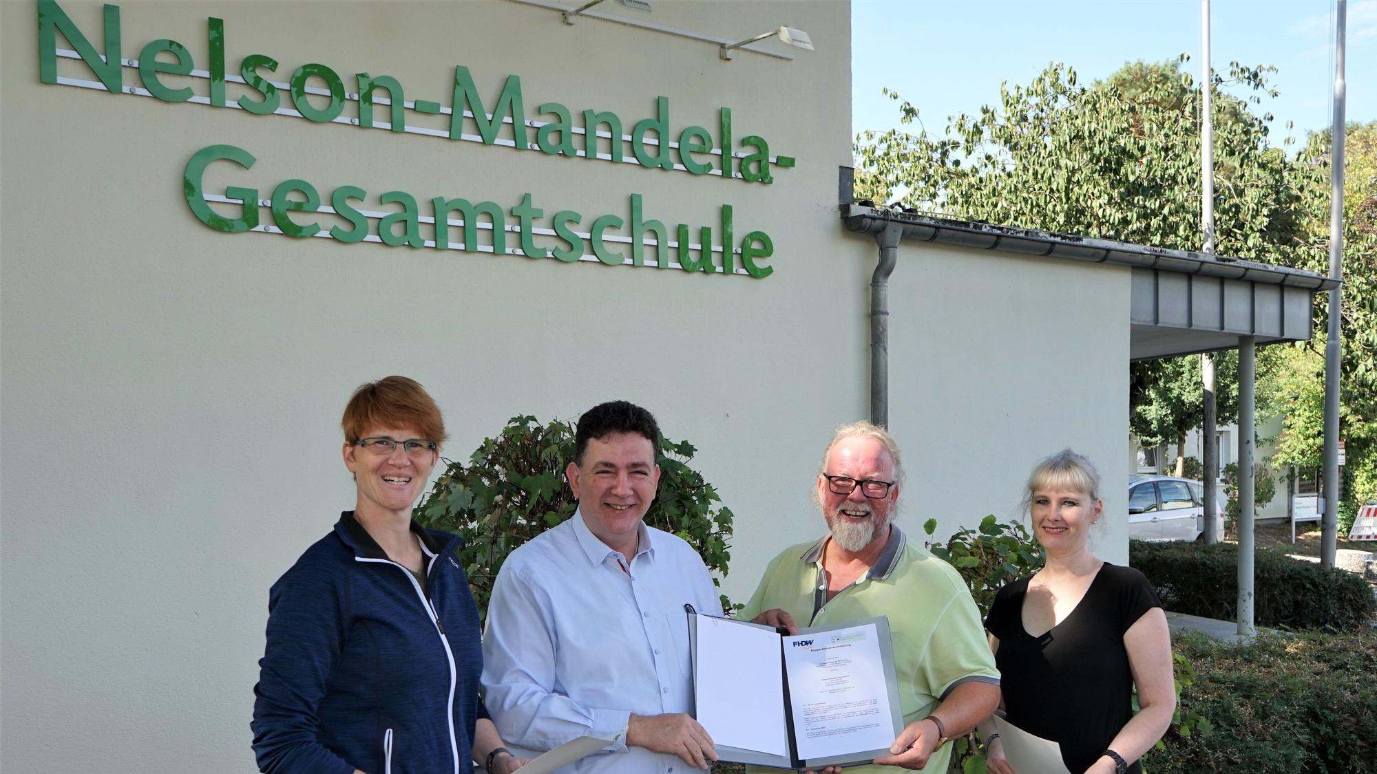 Vier Personen halten einen Vertrag vor dem Gebäude der Nelson-Mandela-Gesamtschule in die Kamera.