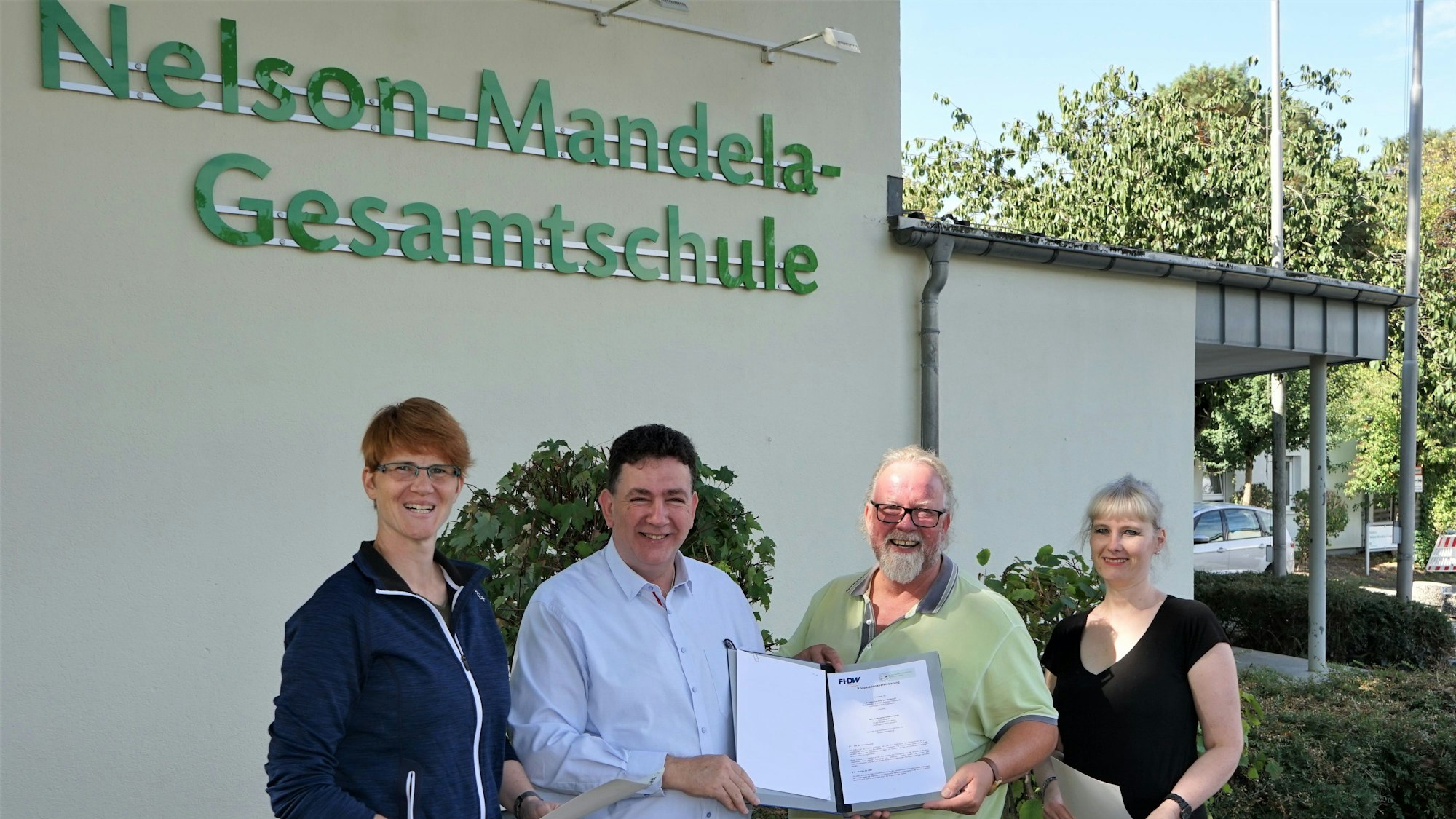 Vier Personen halten einen Vertrag vor dem Gebäude der Nelson-Mandela-Gesamtschule in die Kamera.