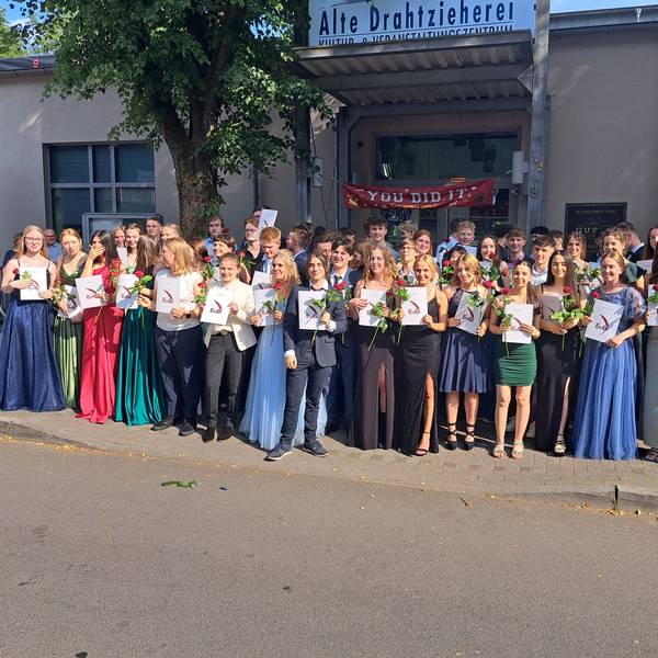 Die Schüler und Schülerinnen stehen vor der Alten Drahtzieherei und lächeln, mit Zeugnis und Rose in der Hand in die Kamera.