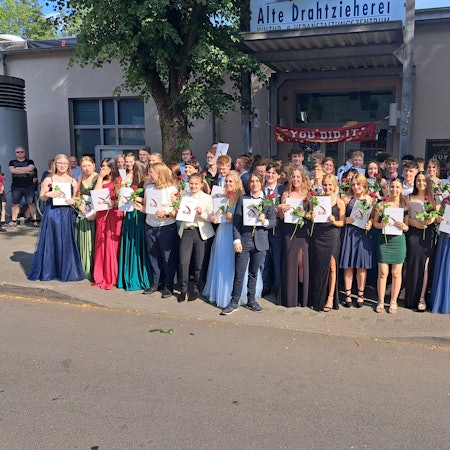 Die Schüler und Schülerinnen stehen vor der Alten Drahtzieherei und lächeln, mit Zeugnis und Rose in der Hand in die Kamera.