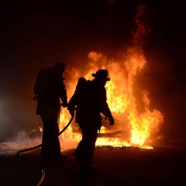 Dunkle Umrisse von zwei Feuerwehrleuten. Im Hintergrund sieht man ein brennendes Autowrack. (Archivbild)