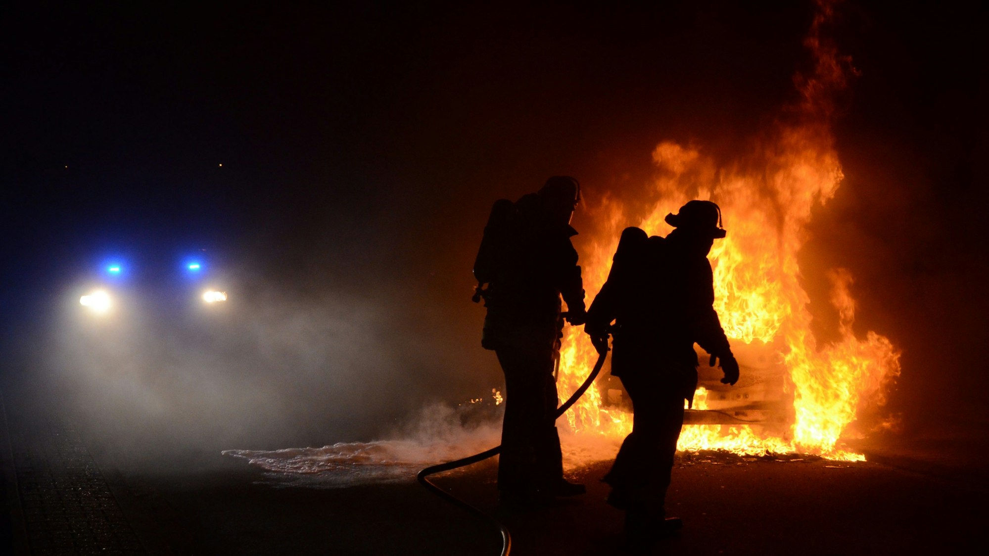 Dunkle Umrisse von zwei Feuerwehrleuten. Im Hintergrund sieht man ein brennendes Autowrack. (Archivbild)