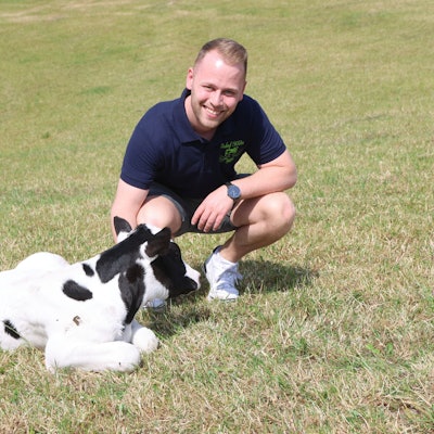 Ein junger Mann hockt auf einer Wiese neben einem Kälbchen.