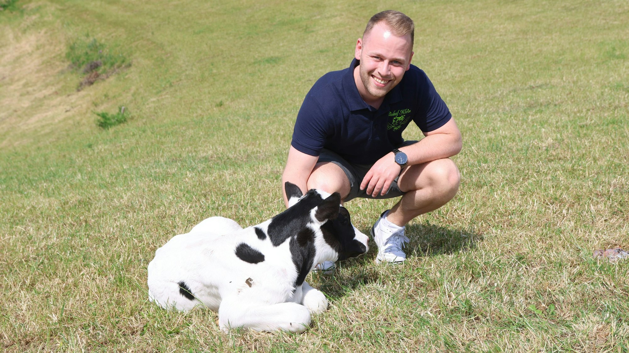 Ein junger Mann hockt auf einer Wiese neben einem Kälbchen.