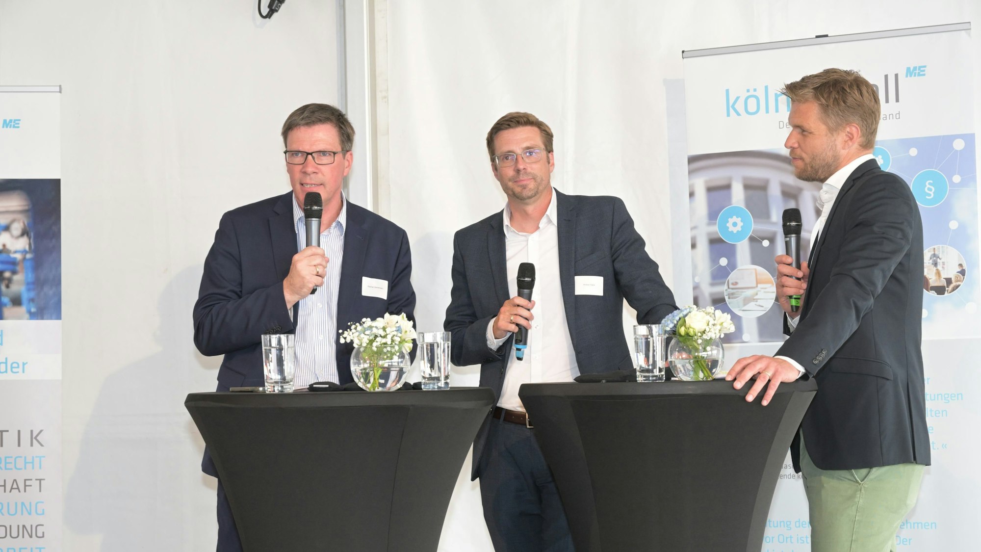 Die Männer stehen auf der Bühne an zwei Stehtischen und halten Mikros in der Hand.