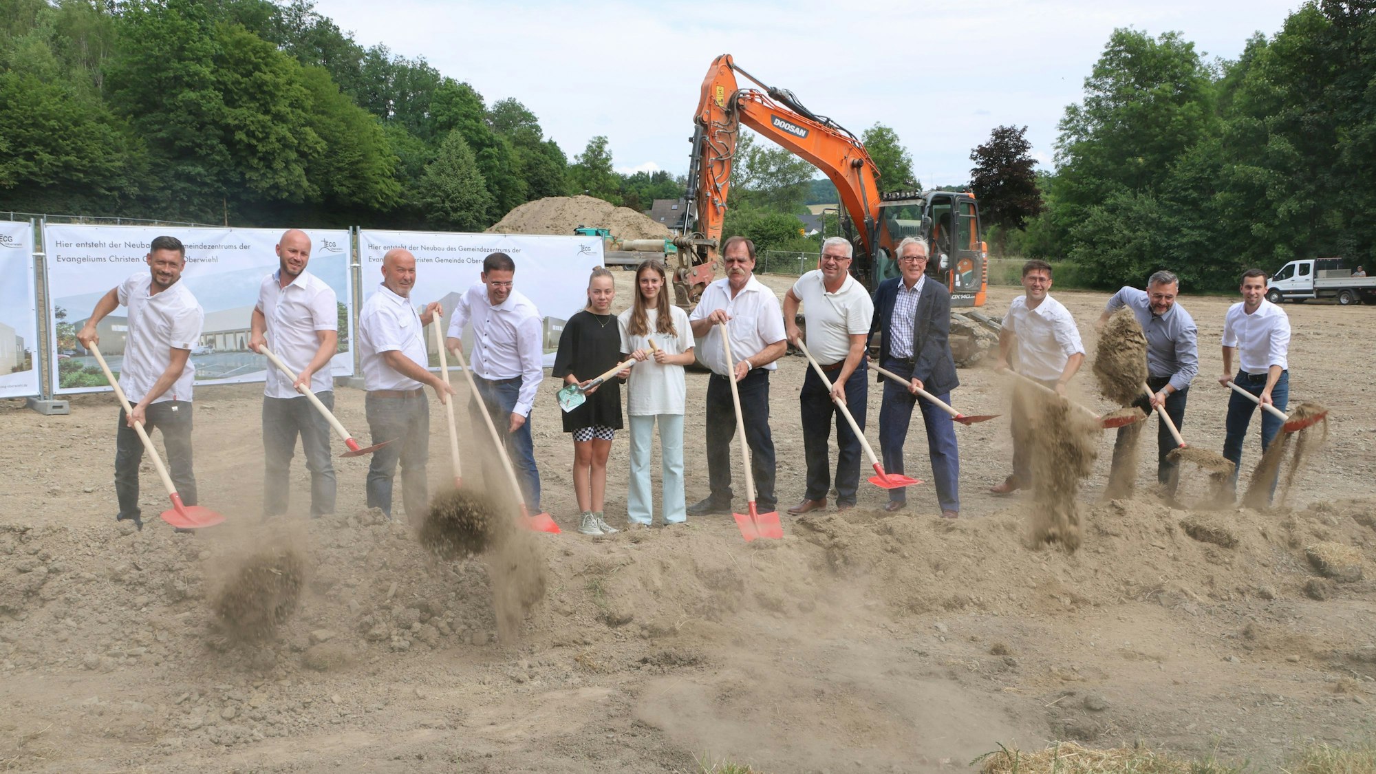 Zwölf Personen stehen vor einem Bagger und werfen mit Schaufeln Erde in die Luft.