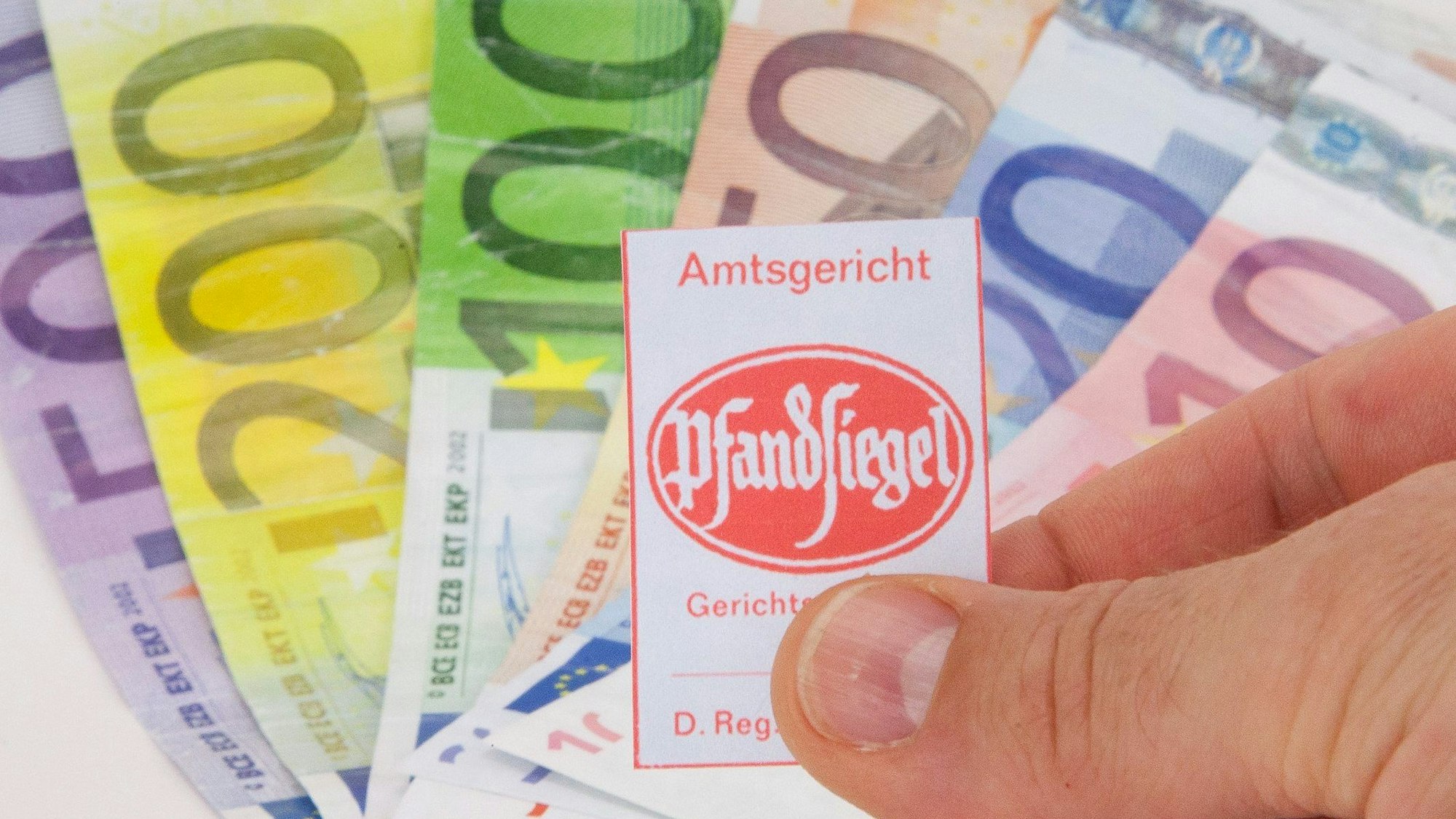 Ein Bündel mit Geldscheinen liegt im Hintergrund, im Vordergrund hält ein Mann ein Pfandsiegel in der Hand.