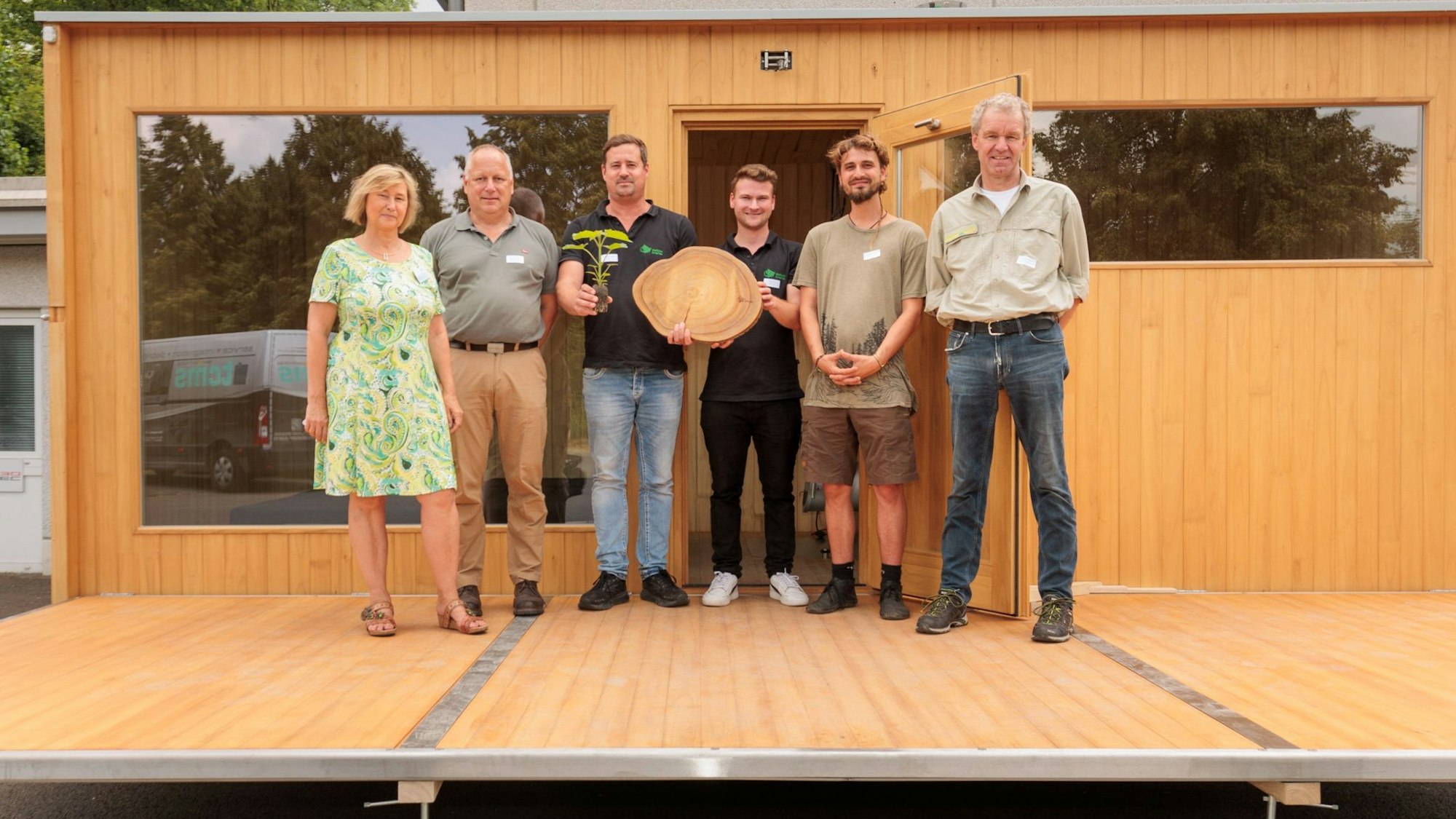 Mehrere Männer und Frauen stehen in einer Reihe. In ihrer Mitte halten sie eine Baumstammscheibe in der Hand.