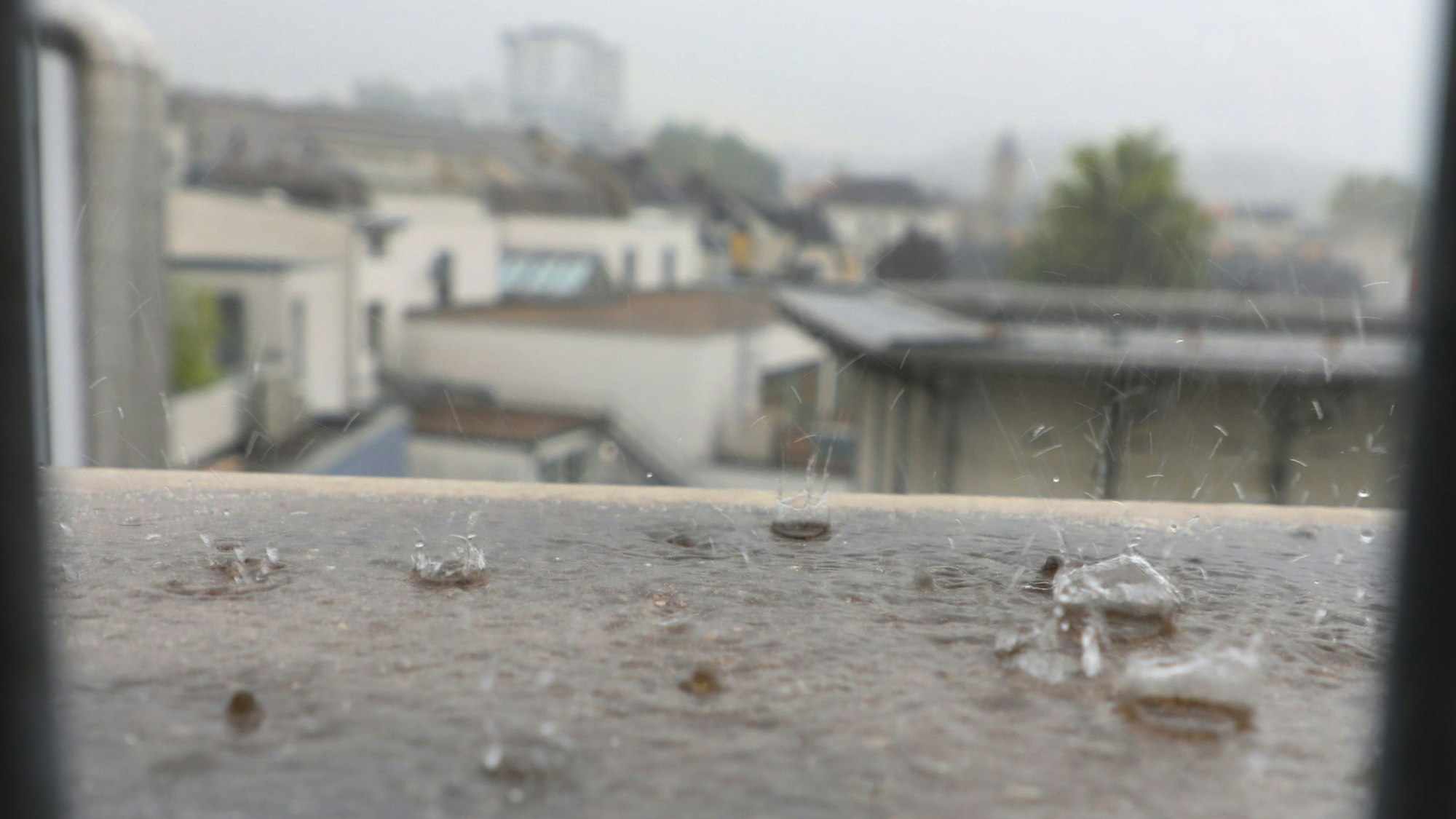 Regentropfen prasseln auf ein Dach, im Hintergrund ist verschwommen die Silhouette von Bergisch Gladbachs Stadtmitte zu sehen.