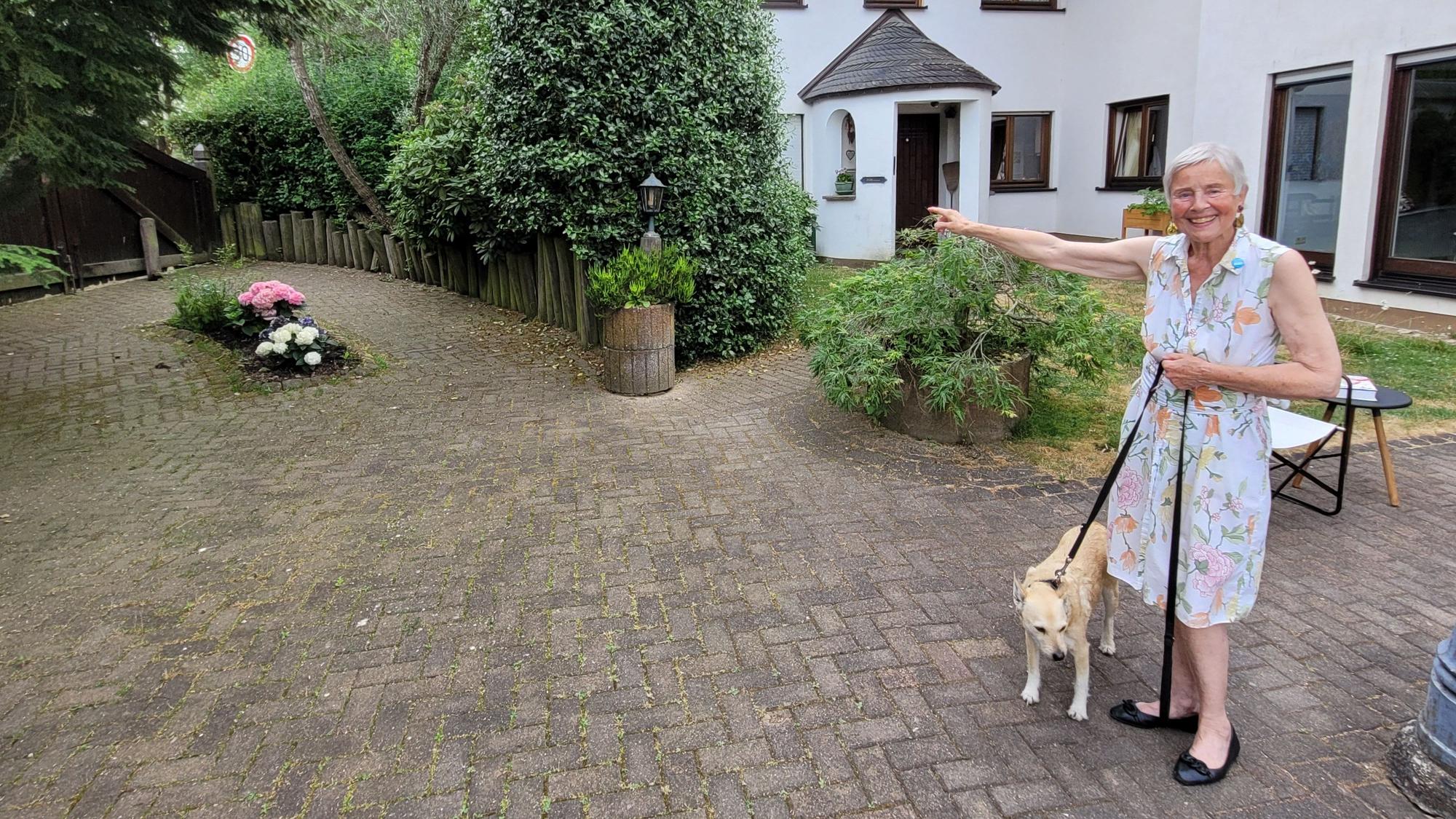 Eine Frau in einem Sommerkleid steht im Hof ihres Hauses und hält einen Hund an der Leine.