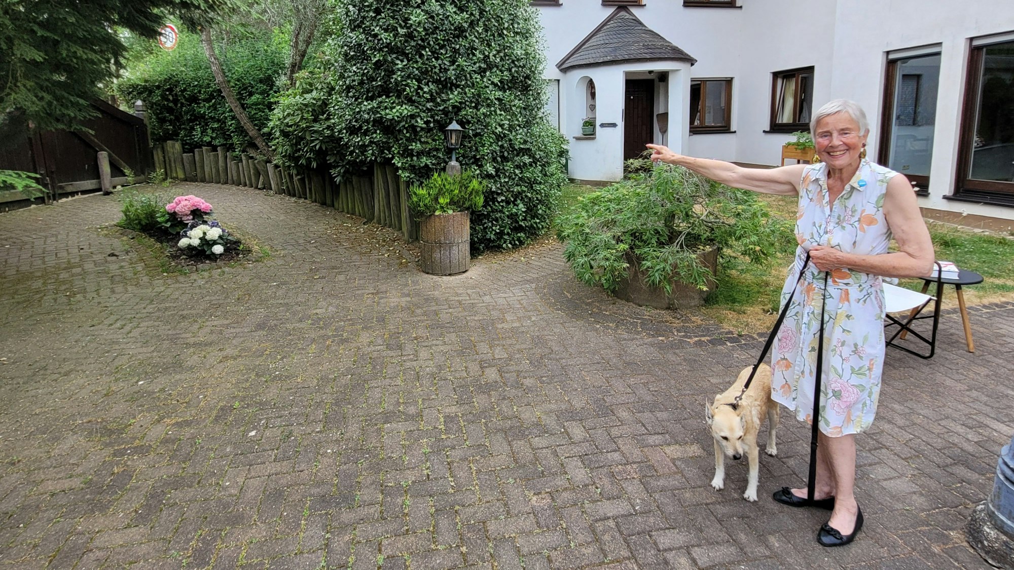 Eine Frau in einem Sommerkleid steht im Hof ihres Hauses und hält einen Hund an der Leine.