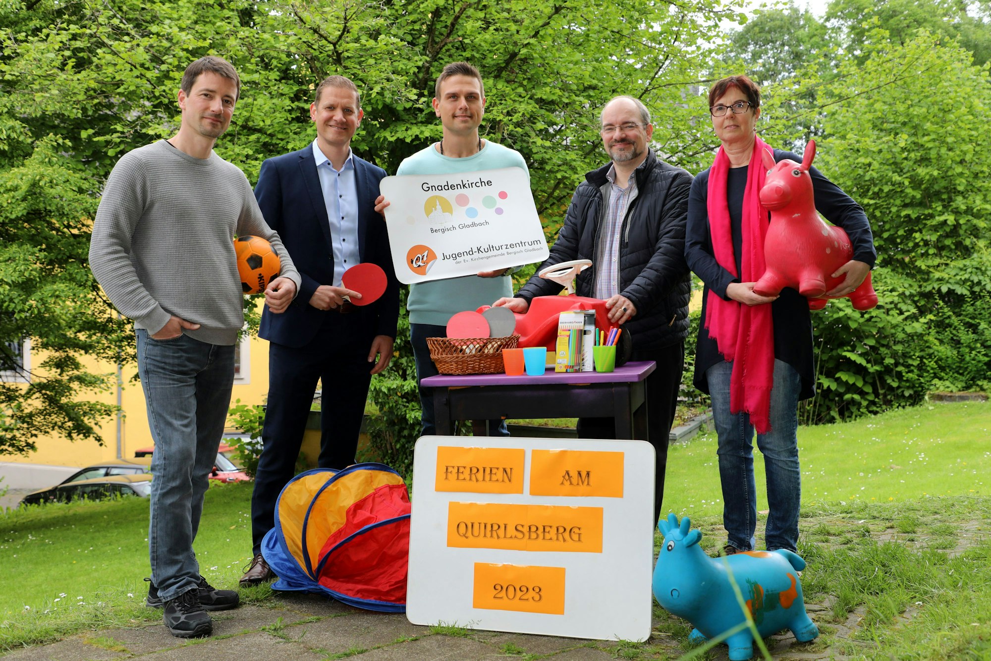 Fünf Menschen stehen mit Spielgeräten und einem Schild „Ferien am Quirlsberg 2023“ auf einer Wiese.